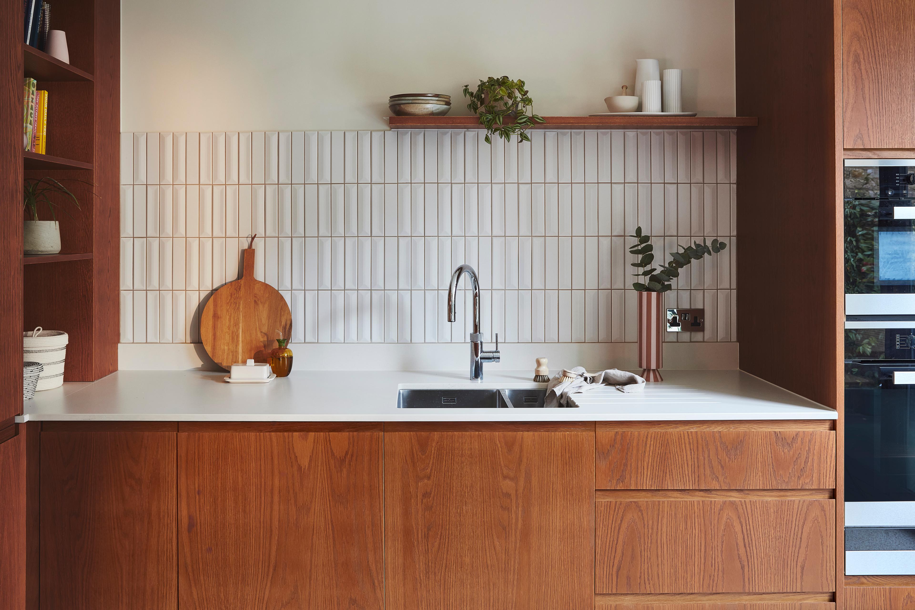 a contemporary kitchen features warm wood cabinetry, a white countertop with an integrated stainless steel sink and chrome faucet, and a vertically stacked white tile backsplash accented by minimalist decor.