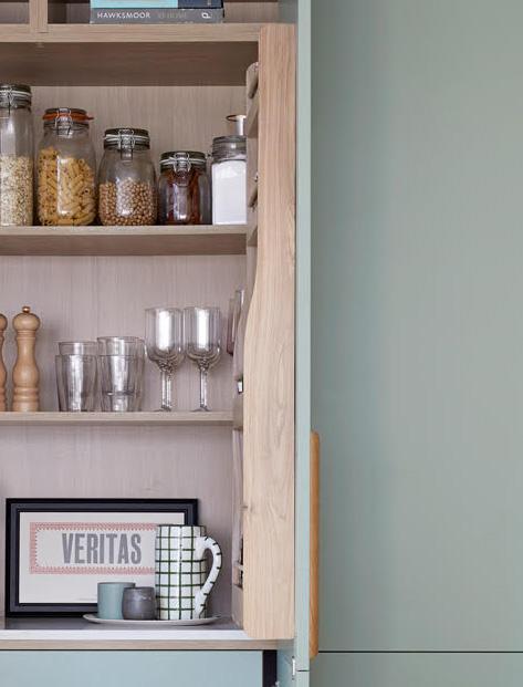 Open kitchen cabinet with shelves holding jars of dry food, drinking glasses, a framed VERITAS print, a mug, and a pepper grinder. The cabinet is pale wood with a light green door.