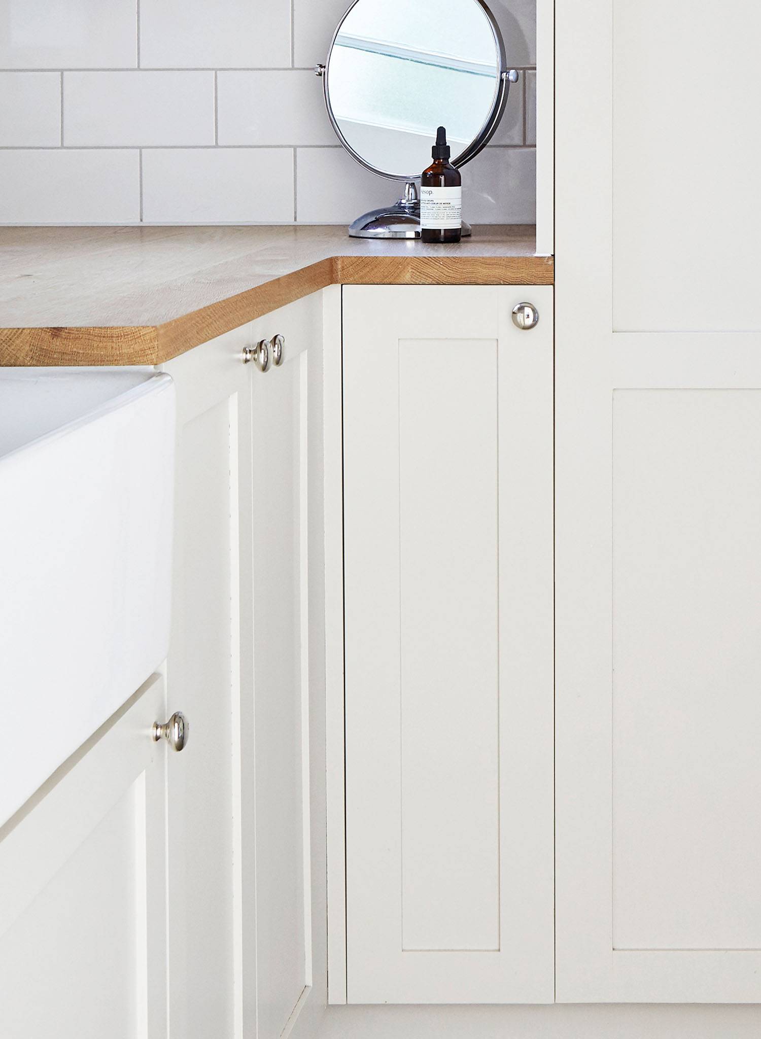 A modern white kitchen corner with shaker-style cabinets, wooden countertop, a round mirror, a bottle, and white subway tile backsplash.