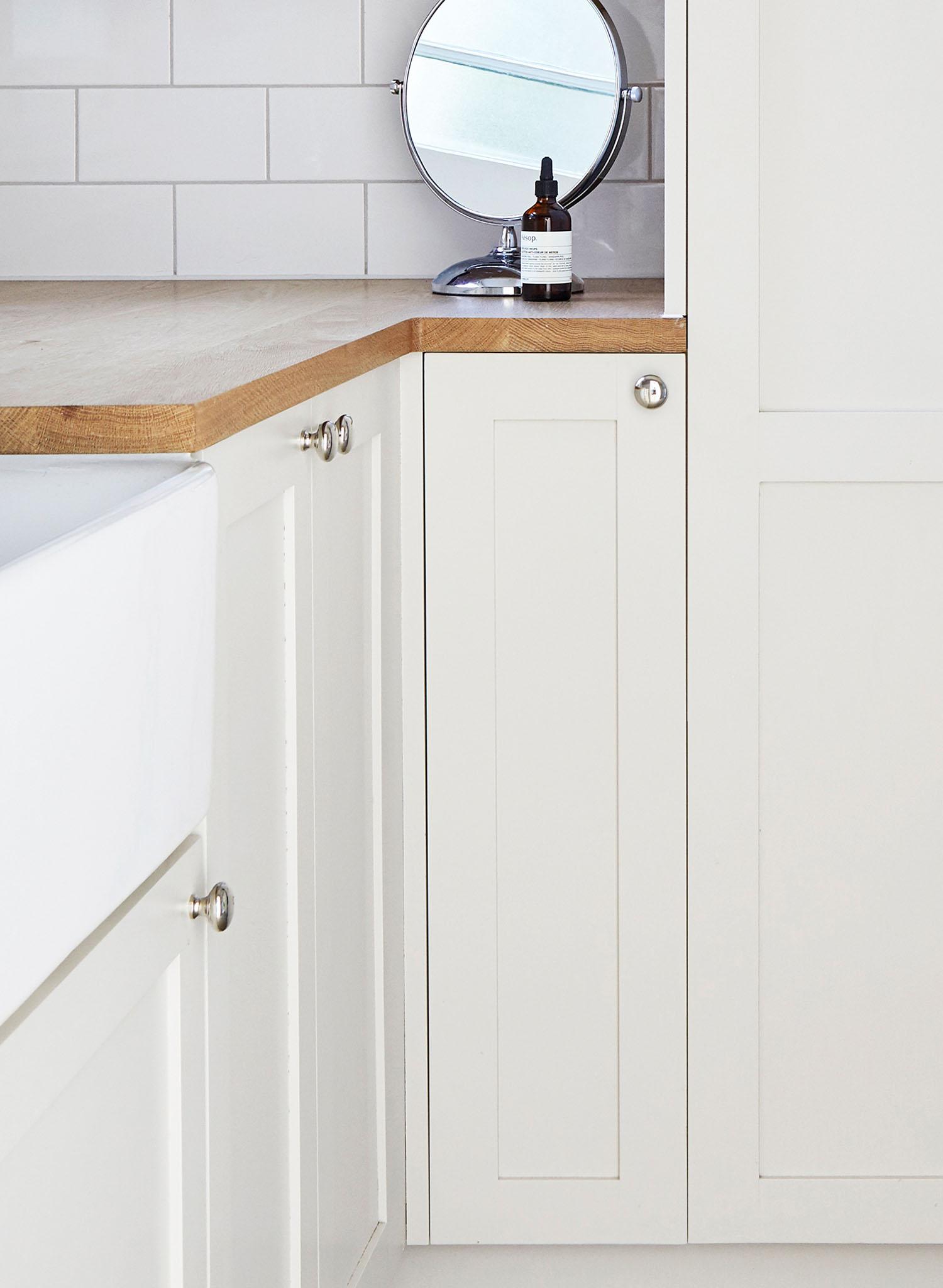 A modern white kitchen corner with shaker-style cabinets, wooden countertop, a round mirror, a bottle, and white subway tile backsplash.