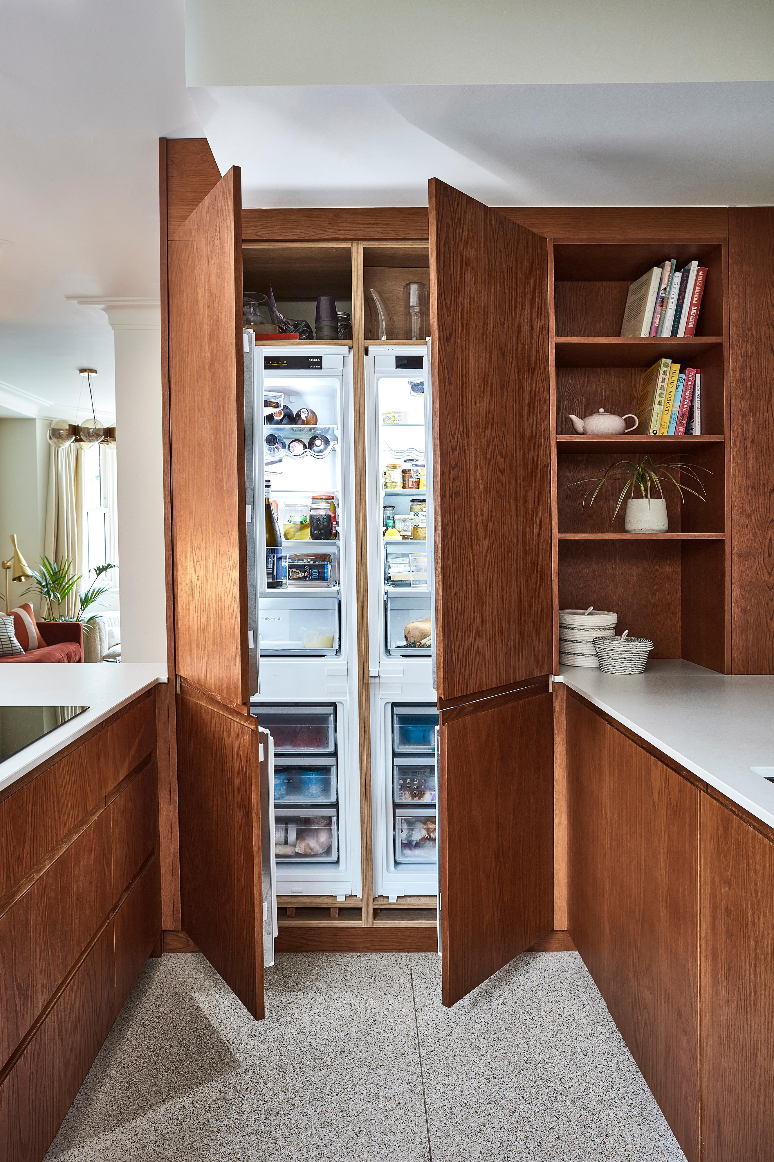 Modern kitchen with sleek wooden cabinets and white countertops. Open shelving displays books and a plant. Natural light streams in through a window, creating a warm, inviting atmosphere.