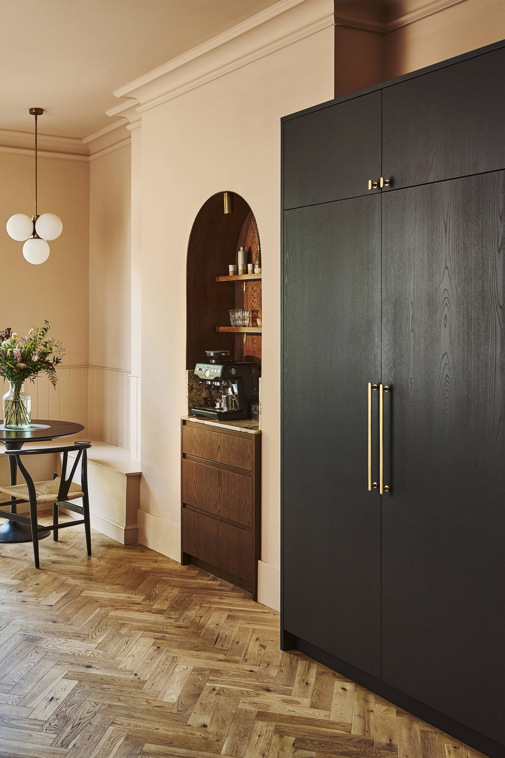A modern kitchen with wooden cabinets, marble backsplash, a built-in oven, cookware on the stove, open shelves holding plates, bowls, framed art, and decorative items, lit by two gold pendant lights.