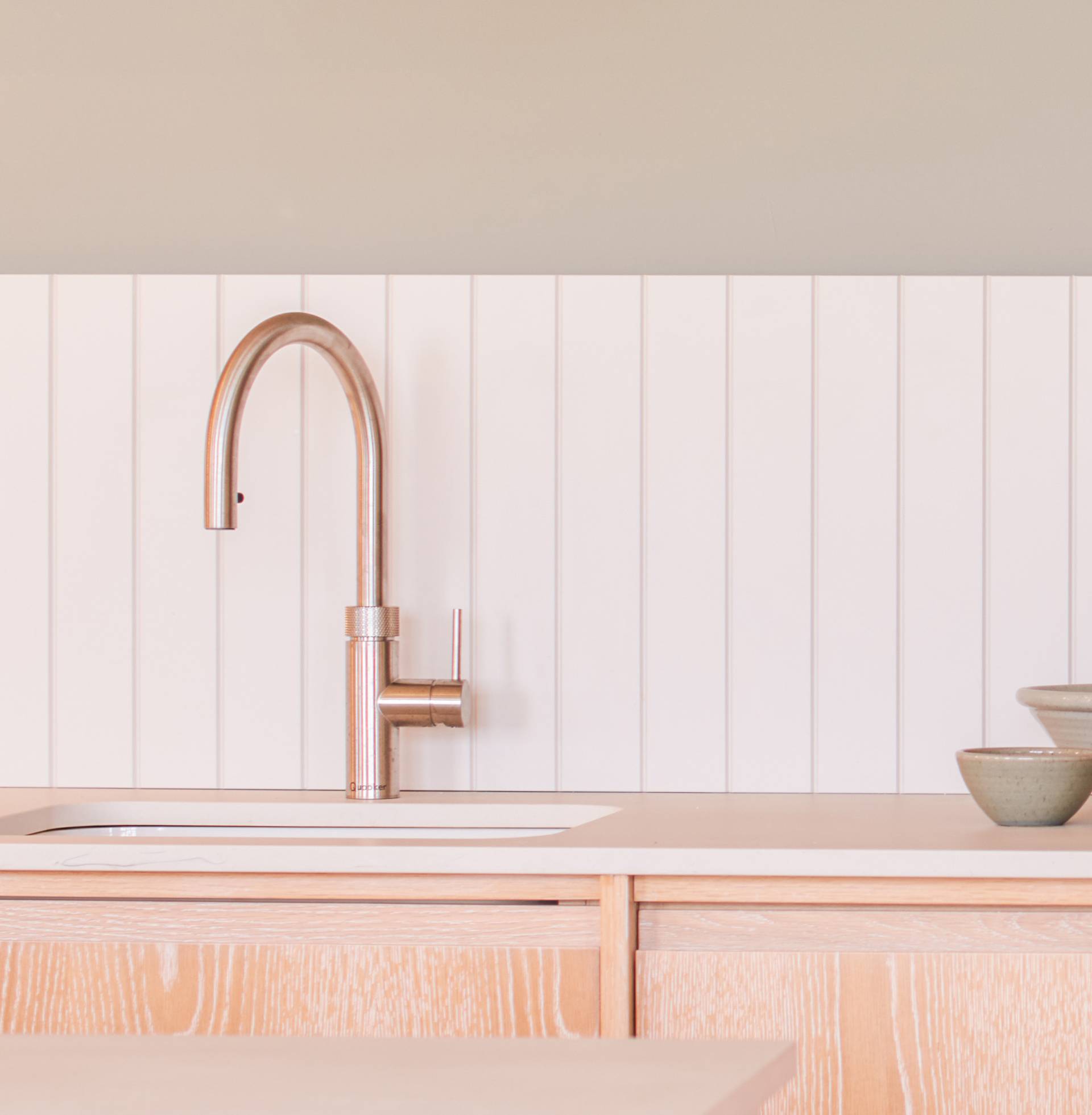 Modern kitchen sink with a sleek, curved faucet set against a white paneled backsplash. The light wood countertop holds two stacked ceramic bowls to the right, creating a minimalist and clean aesthetic.