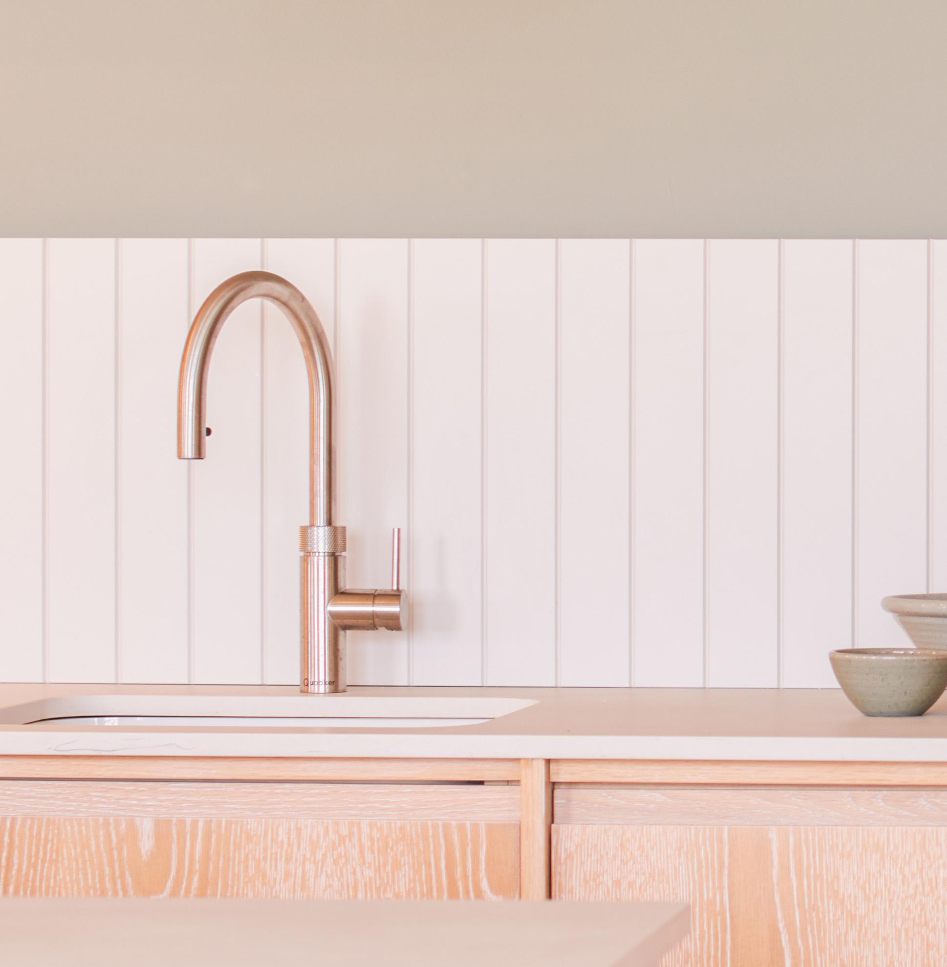 Modern kitchen sink with a sleek, curved faucet set against a white paneled backsplash. The light wood countertop holds two stacked ceramic bowls to the right, creating a minimalist and clean aesthetic.