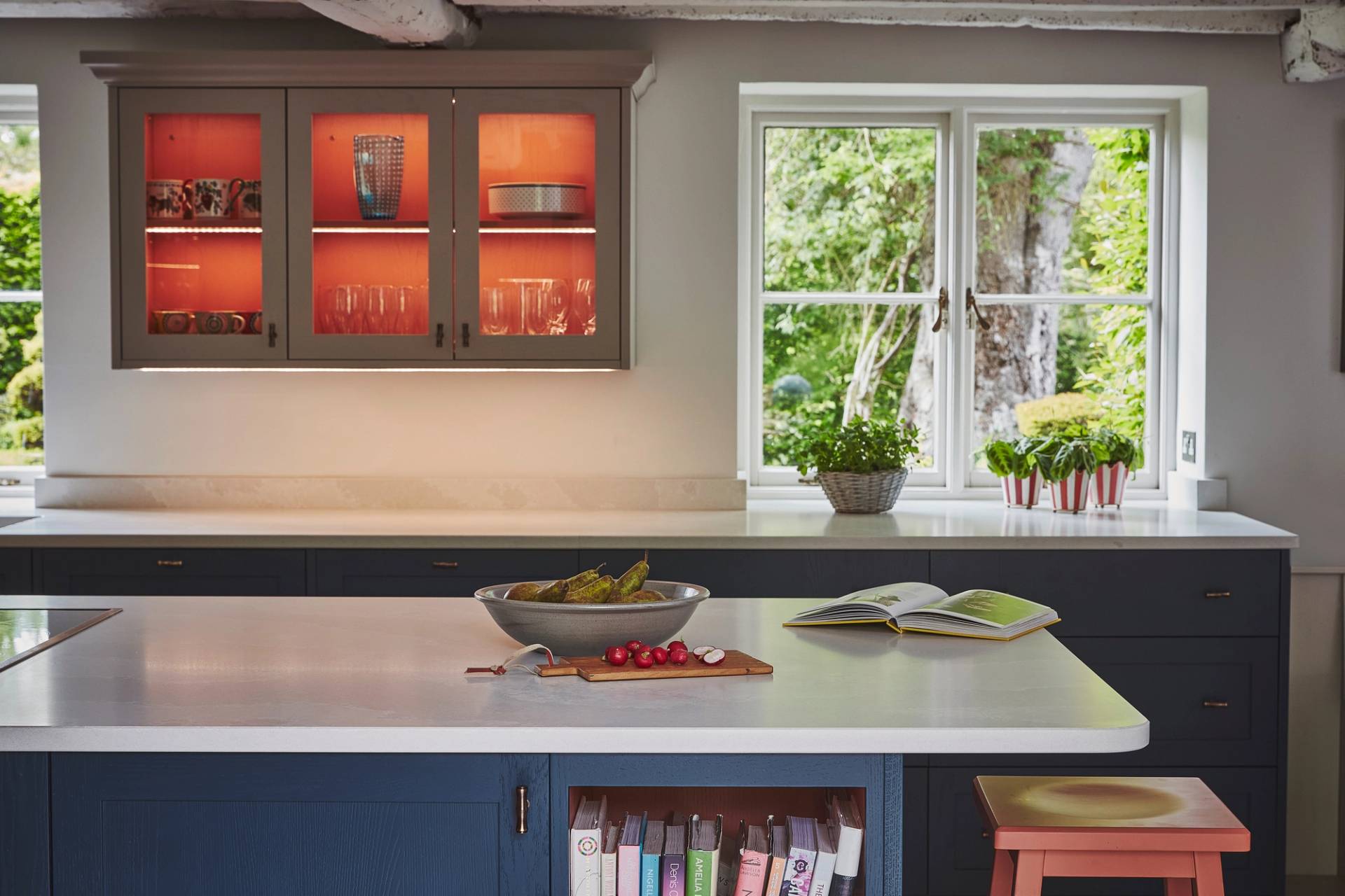 A modern kitchen with navy blue cabinets, a white countertop, cookbooks on a shelf, a bowl of fruit, an open cookbook, radishes on a cutting board, a window with greenery outside, and lit glass cabinets.