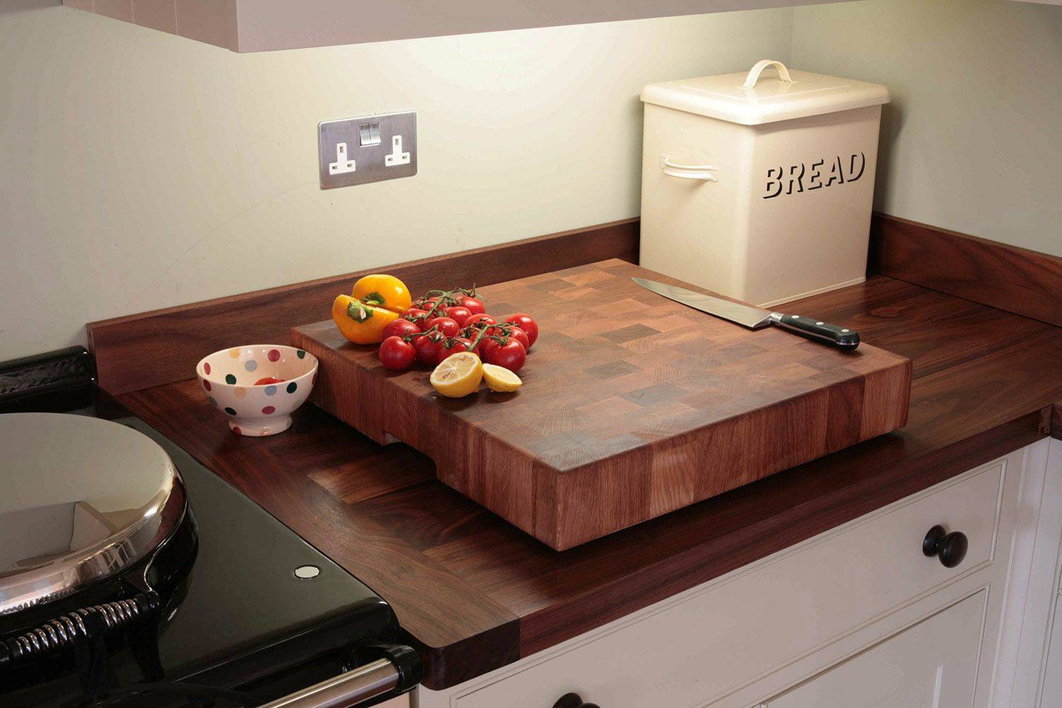 A kitchen countertop with a large wooden cutting board. On the board are tomatoes, a yellow bell pepper, and a sliced lemon. A knife rests nearby. There's a polka-dotted bowl and a cream-colored bread box in the background.