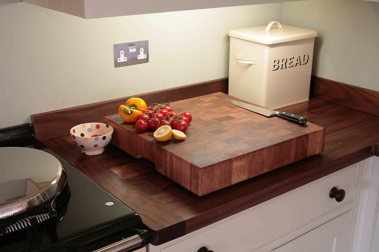 A kitchen countertop with a large wooden cutting board. On the board are tomatoes, a yellow bell pepper, and a sliced lemon. A knife rests nearby. There's a polka-dotted bowl and a cream-colored bread box in the background.