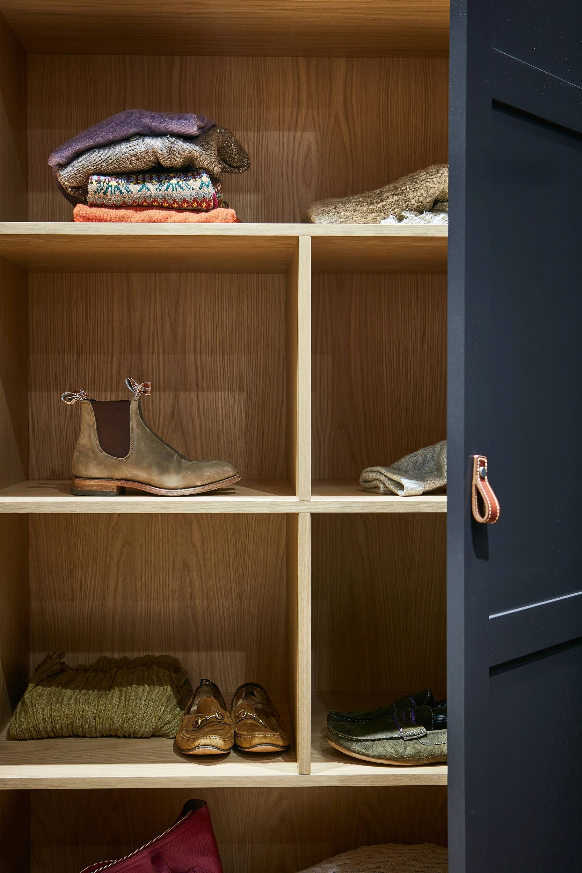 A wooden closet with six shelves, holding folded sweaters, shoes, and a pair of boots. The closet door is partially open, showing a leather loop handle.
