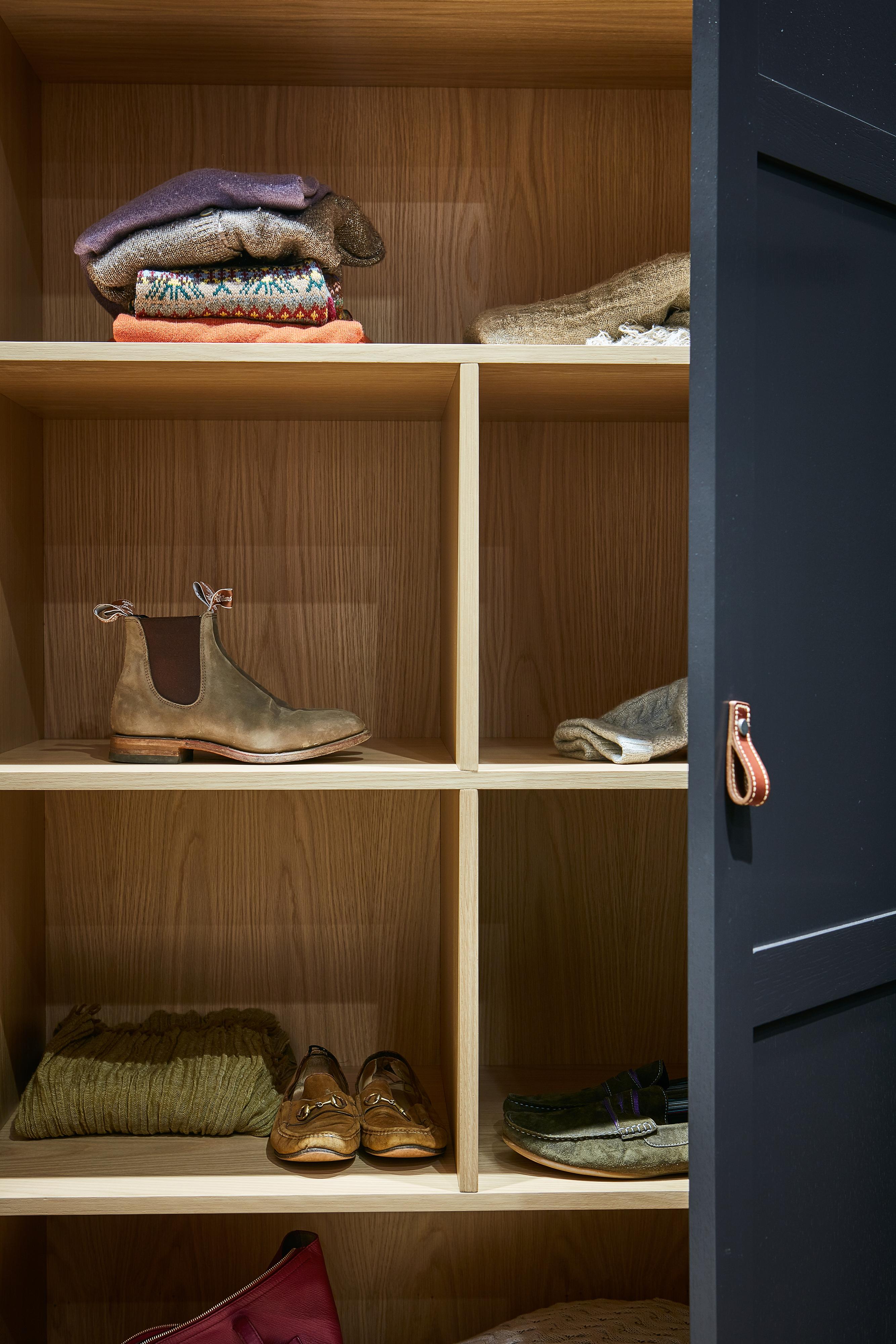 A wooden closet with six shelves, holding folded sweaters, shoes, and a pair of boots. The closet door is partially open, showing a leather loop handle.