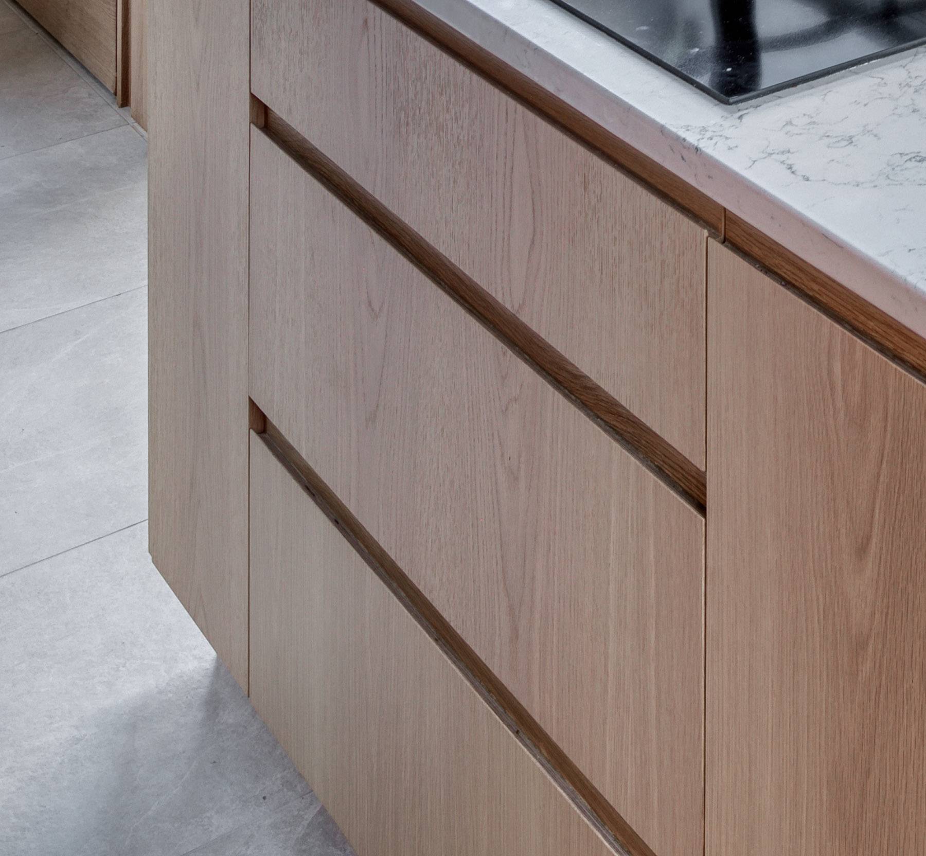 Light wood kitchen cabinets with a minimalist design, featuring no visible handles. The countertop above is white with gray veining, and a portion of a black cooktop is visible. The floor is light gray tile.