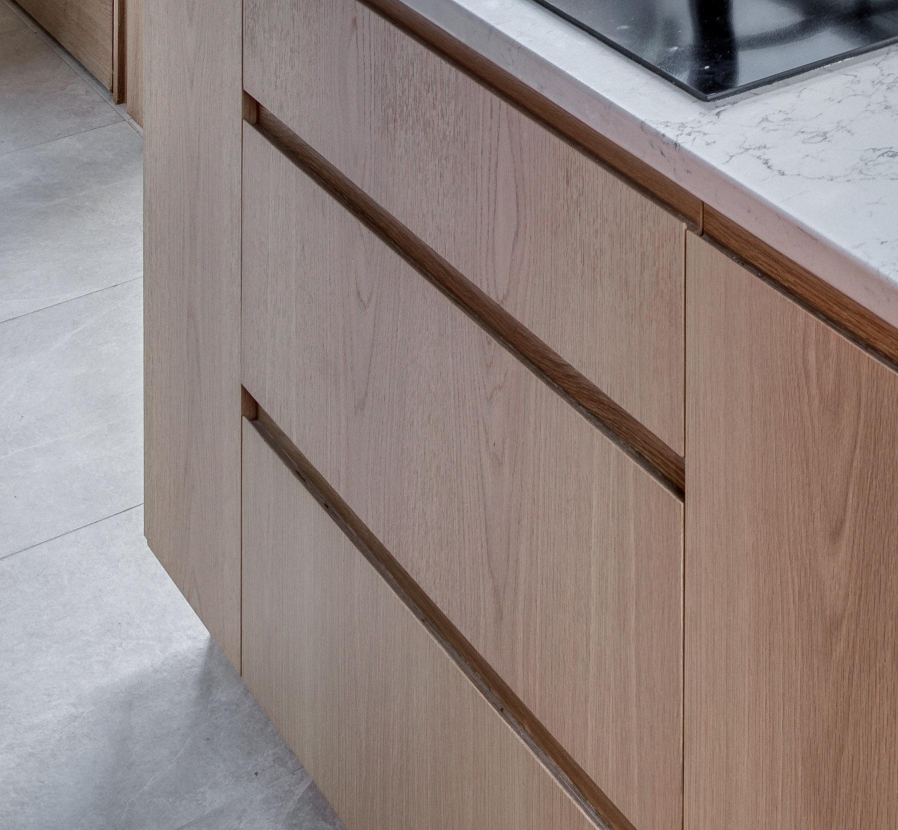 Light wood kitchen cabinets with a minimalist design, featuring no visible handles. The countertop above is white with gray veining, and a portion of a black cooktop is visible. The floor is light gray tile.