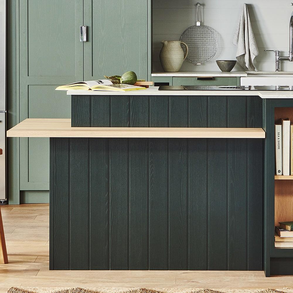 Modern kitchen with a green island featuring a light wood countertop extension. The background has green cabinets, a sink, a woven basket, a folded towel, and a shelf holding books and decorative items.