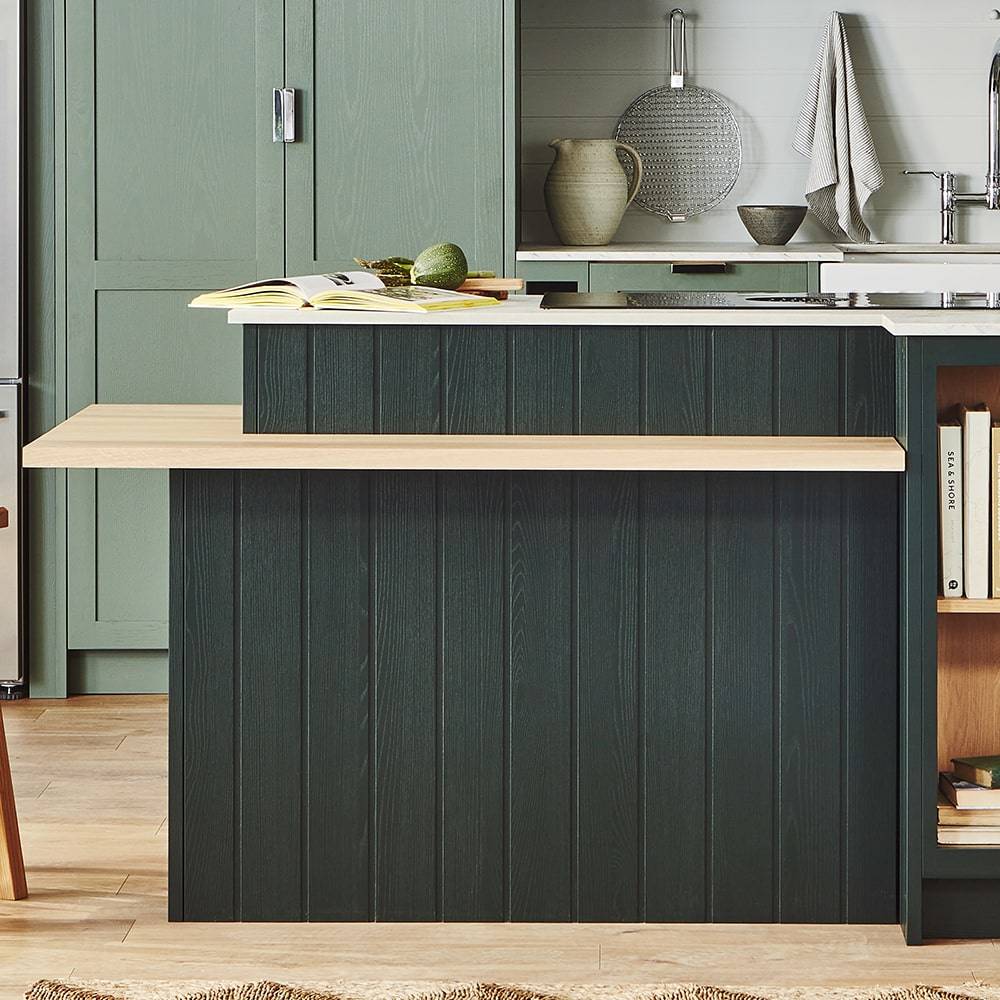 Modern kitchen with a green island featuring a light wood countertop extension. The background has green cabinets, a sink, a woven basket, a folded towel, and a shelf holding books and decorative items.