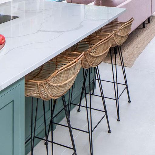 Three wicker barstools with black metal legs are lined up under a light gray marble kitchen island with a pale green base. A beige carpet and part of a pink sofa are visible in the background.