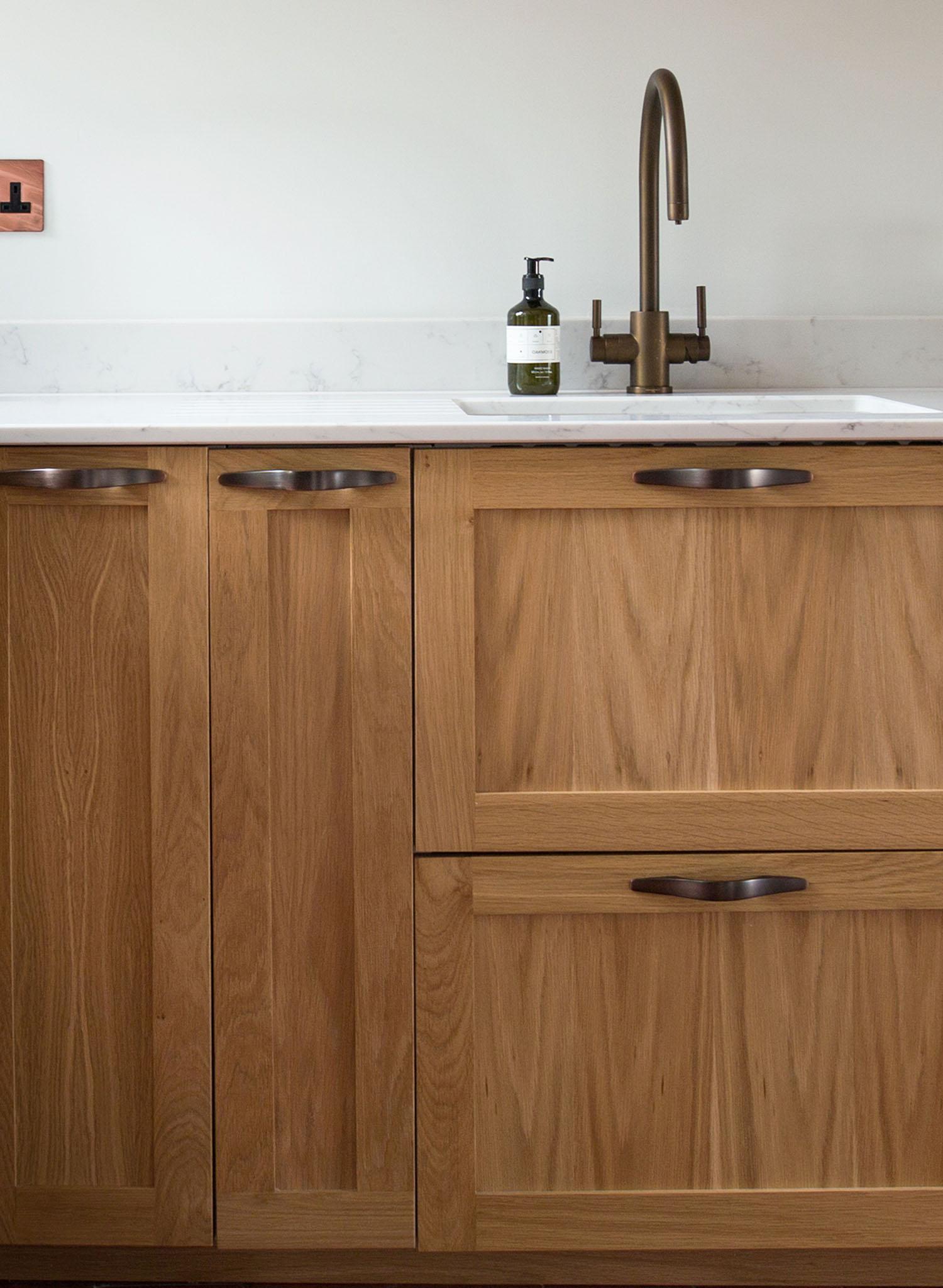 A modern kitchen sink with a bronze faucet, soap dispenser, and natural wood cabinets with sleek handles beneath a white countertop and backsplash.