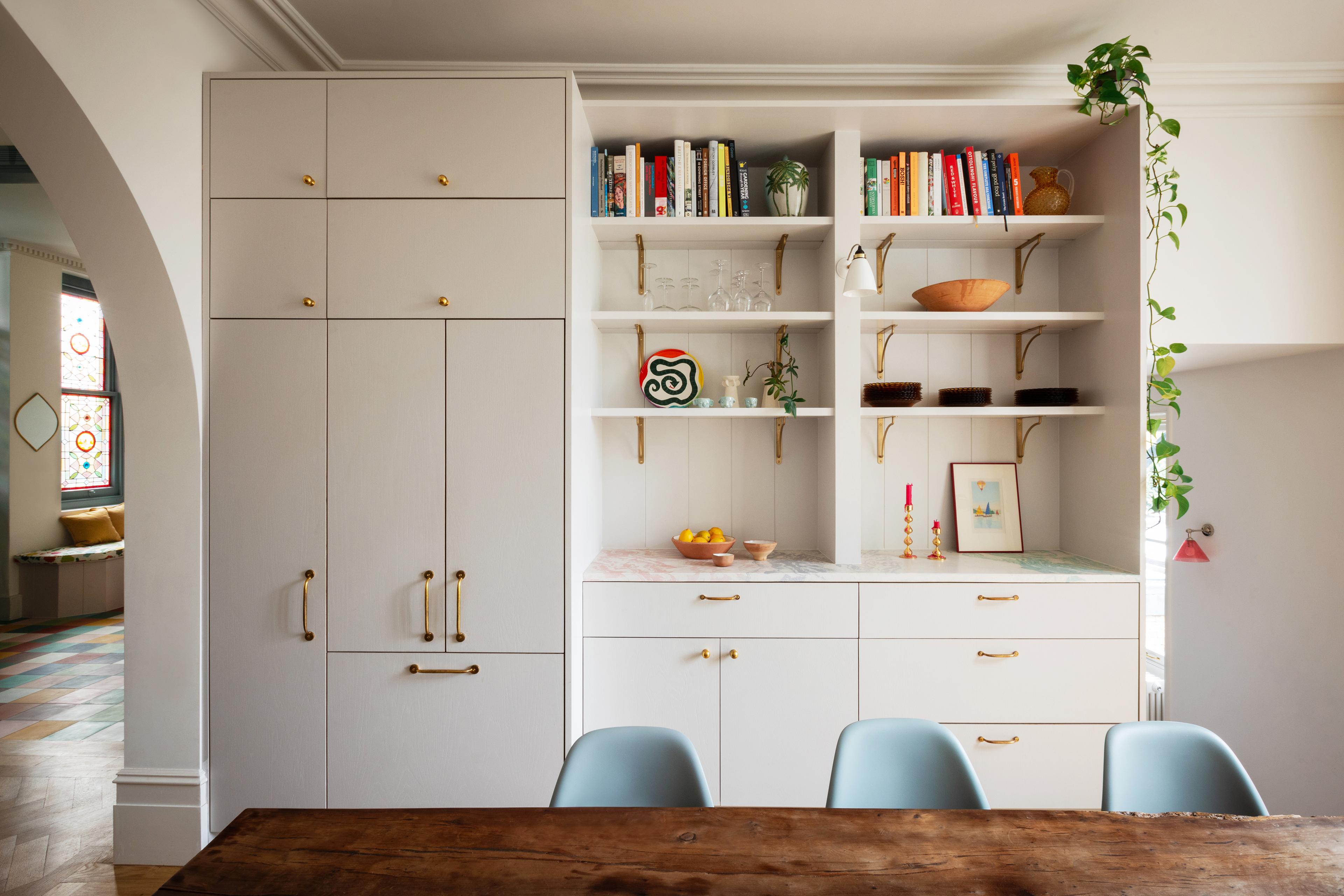 A modern kitchen features light cabinetry with brass handles, open shelves displaying books and decor, a wooden dining table with blue chairs, and a trailing plant hanging from the top shelf.