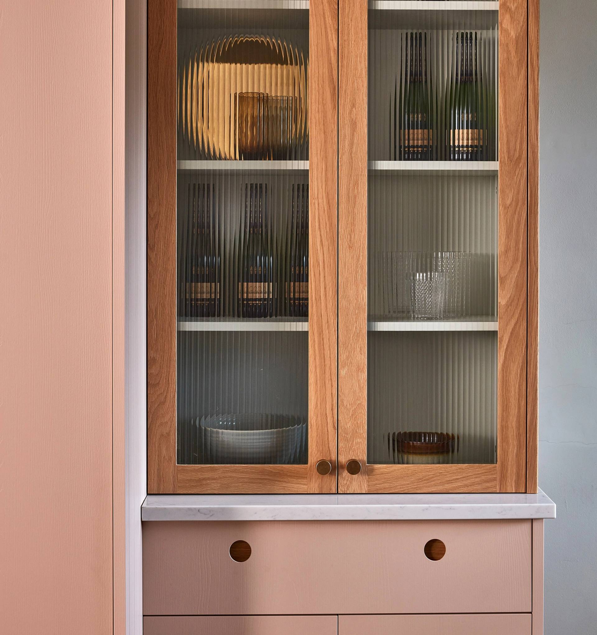 A wooden cabinet with glass doors displays wine bottles, a bowl, and glassware on the shelves. The cabinet has a light marble countertop and two drawers below, set against a soft pink wall