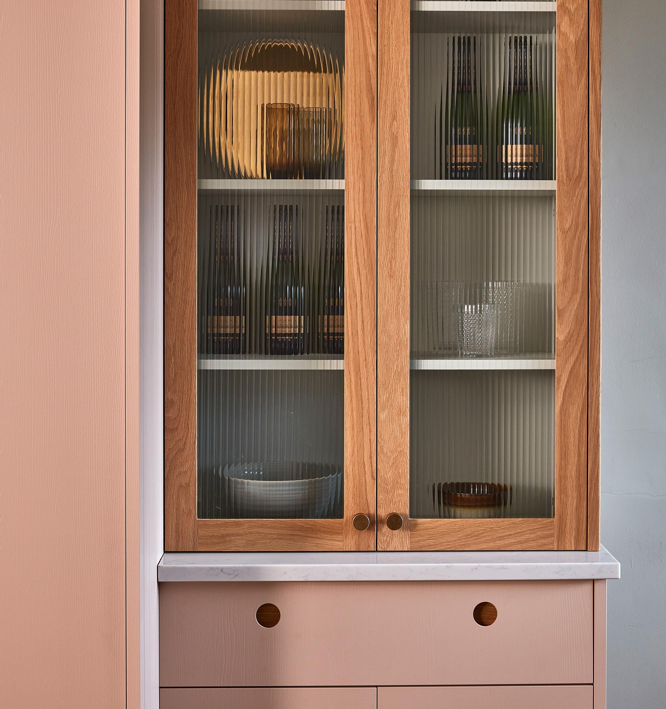 A wooden cabinet with glass doors displays wine bottles, a bowl, and glassware on the shelves. The cabinet has a light marble countertop and two drawers below, set against a soft pink wall
