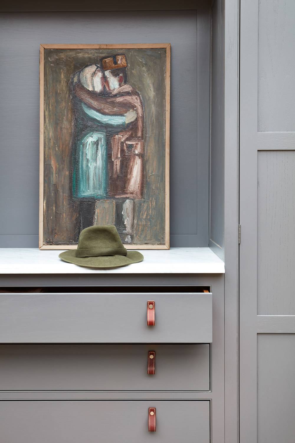 A green felt hat sits on a marble shelf in front of an abstract painting of two people embracing, all set within light gray cabinetry with an open drawer and leather pull handles.