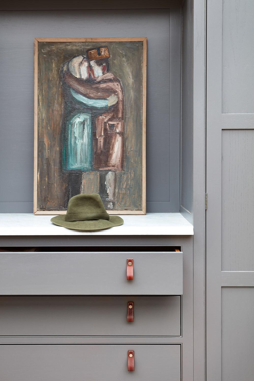 A green felt hat sits on a marble shelf in front of an abstract painting of two people embracing, all set within light gray cabinetry with an open drawer and leather pull handles.