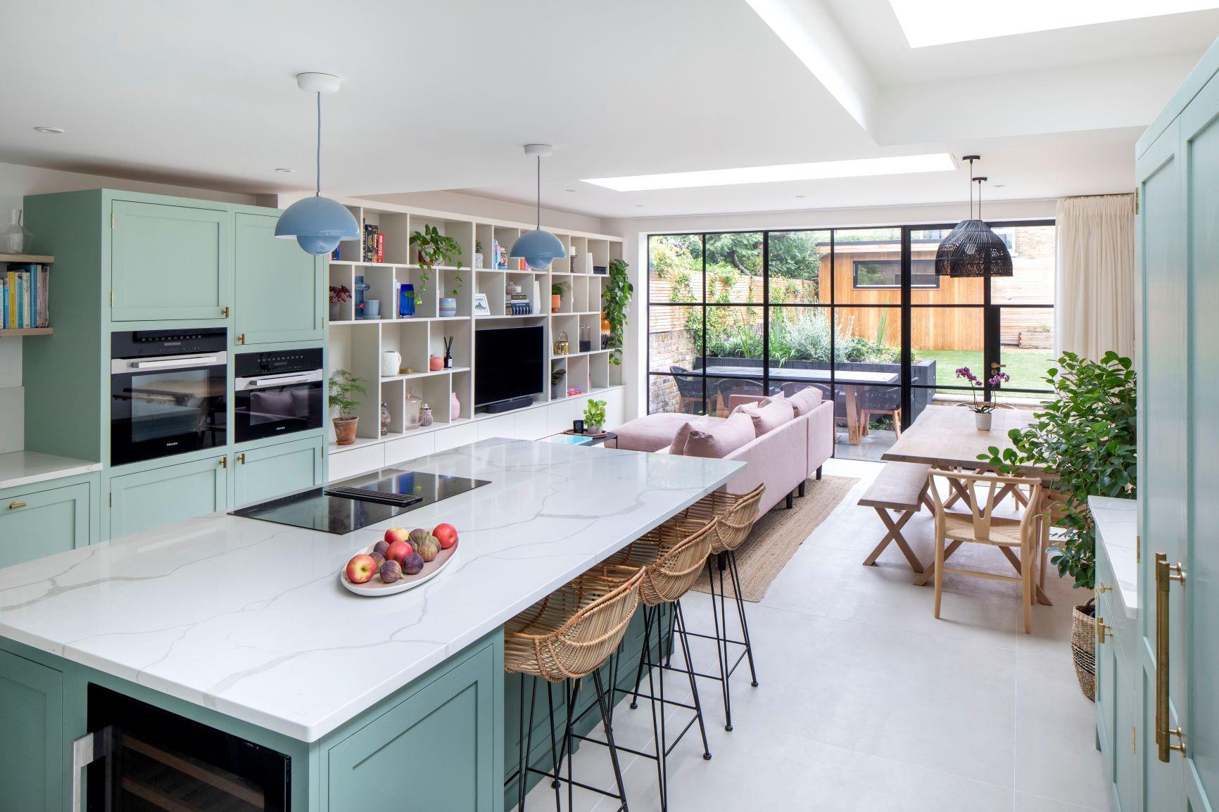 A wide-angle shot of the Dulwich kitchen, highlighting the open-plan layout and the seamless integration between the kitchen and dining area. The light green cabinets create a sense of calm and tranquility, while the marble effect quartz worktops add a touch of luxury.