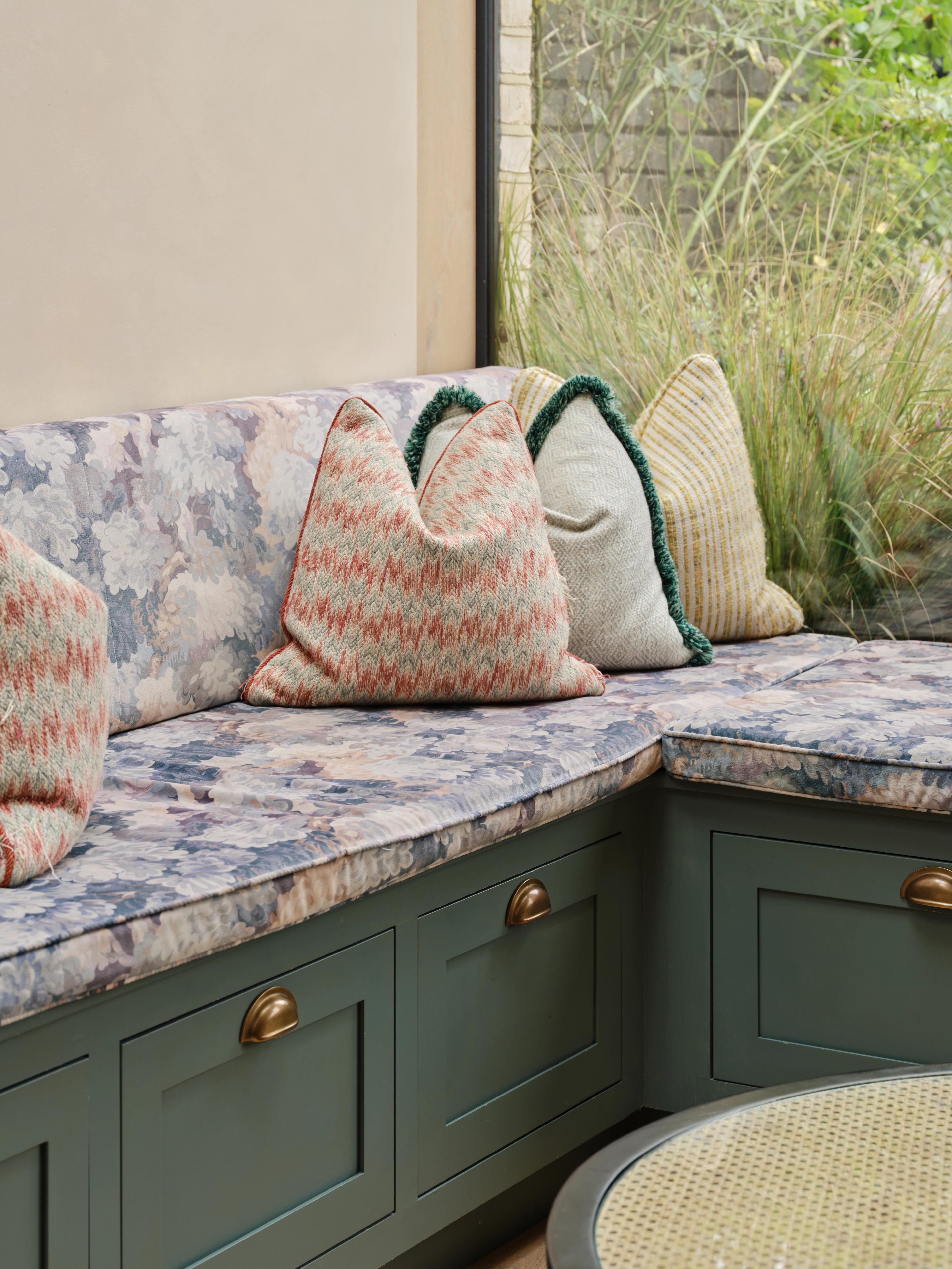 Queens Park kitchen, L-shaped built-in bench seating turning the corner of a window, with dark green in-frame drawer fronts and brass cup handles below a patterned fitted cushion and scatter cushions, and planting visible outside.