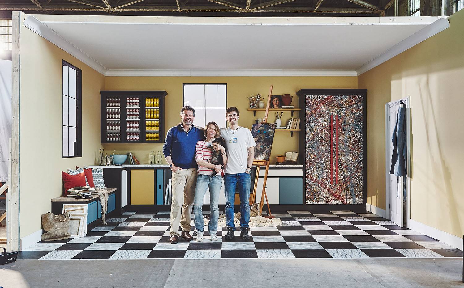 Family stood in kitchen with multicoloured cabinetry and artistic paint splashes covering double cabinet. Walls are yellow, flooring is black and white checkers.