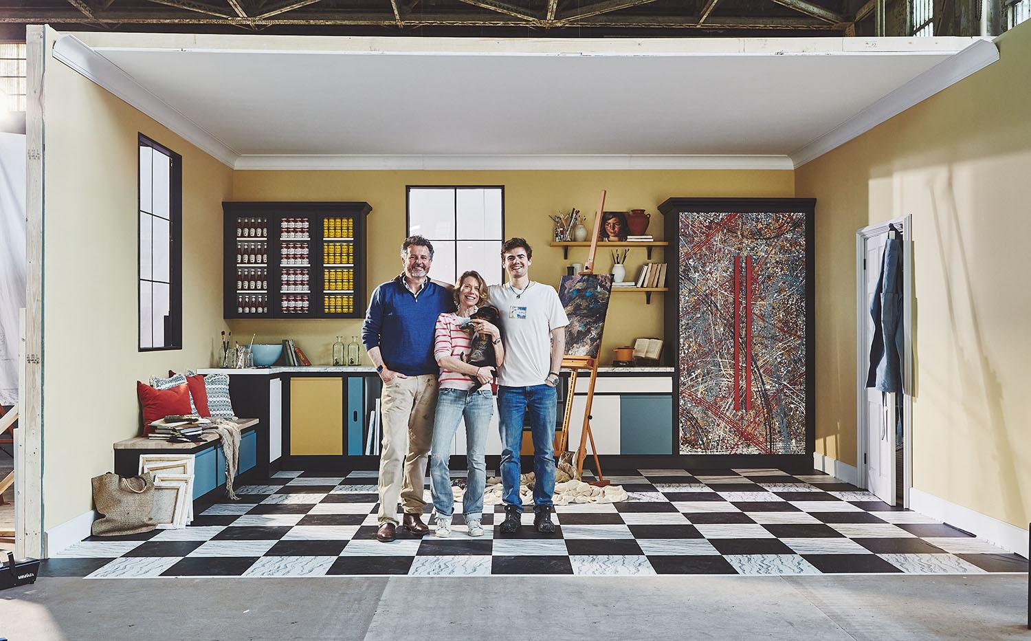 Family stood in kitchen with multicoloured cabinetry and artistic paint splashes covering double cabinet. Walls are yellow, flooring is black and white checkers.
