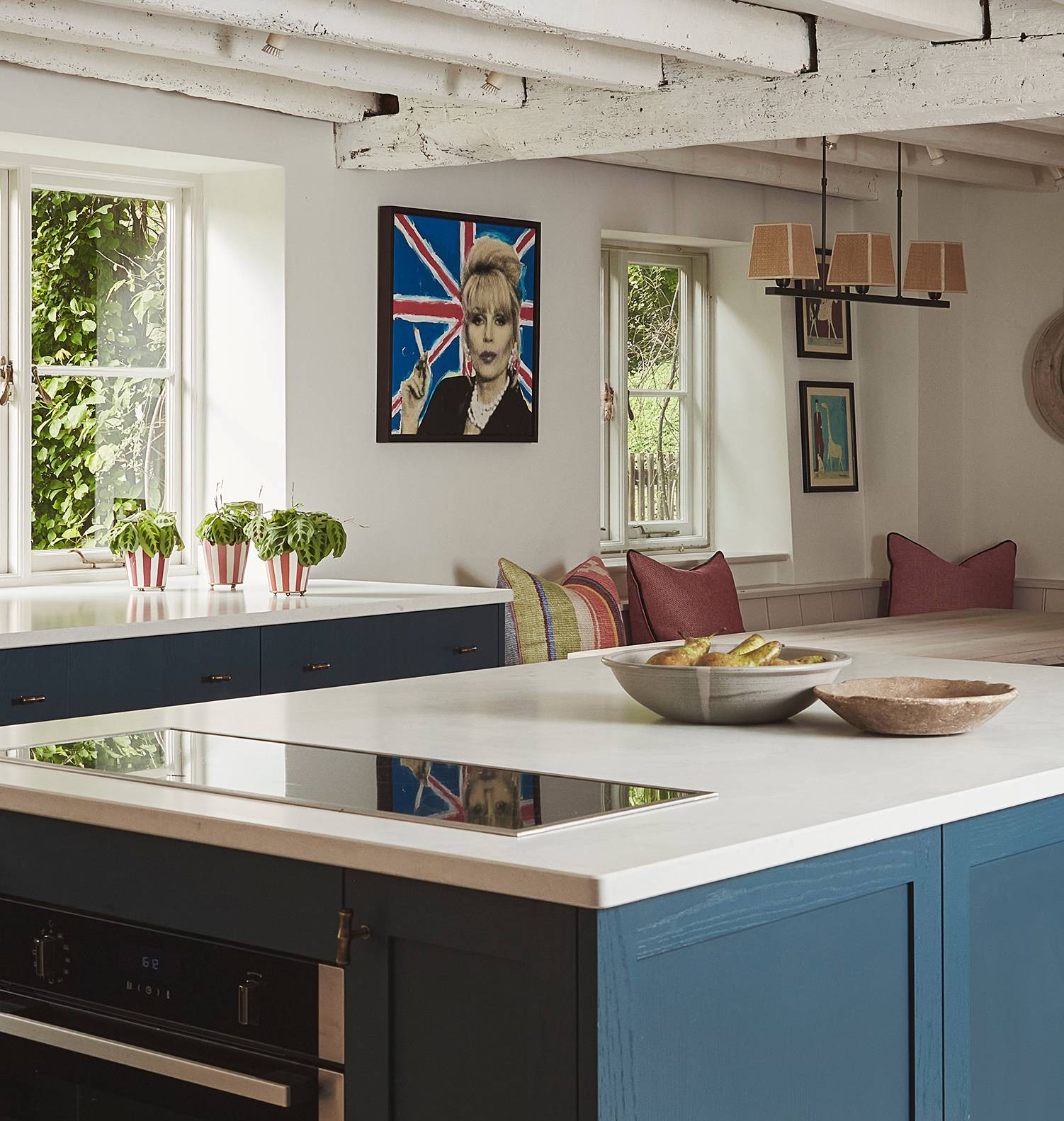 A bright kitchen with blue cabinets, white countertops, potted herbs by the window, a bowl of fruit, and a dining nook with red cushions. A pop art portrait and Union Jack flag hang on the wall. Exposed beams on the ceiling.