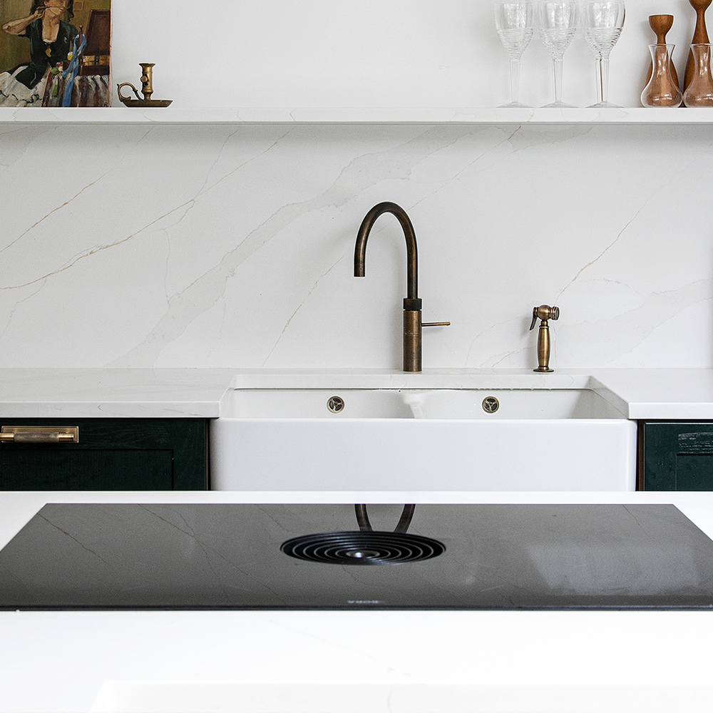 A modern kitchen with a white farmhouse sink, brass faucet, white marble backsplash, and green cabinets. Above, a shelf holds glasses, artwork, and decorative items. In the foreground is a black electric cooktop.