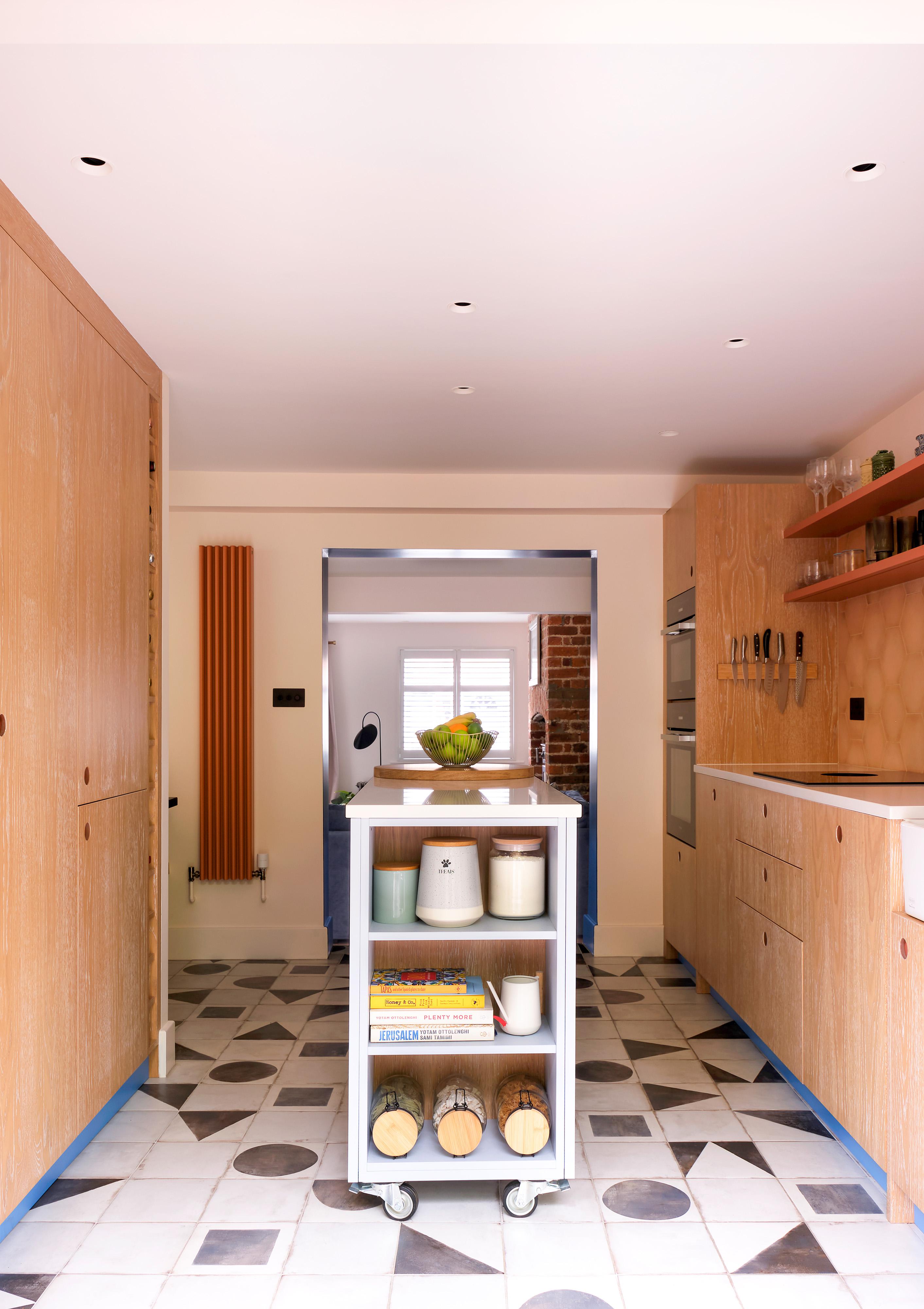 A modern kitchen with light wood cabinets and geometric patterned floor tiles. A rolling cart in the center holds containers and books. The kitchen features a built-in oven, floating shelves with utensils, and a bowl of green apples on the countertop