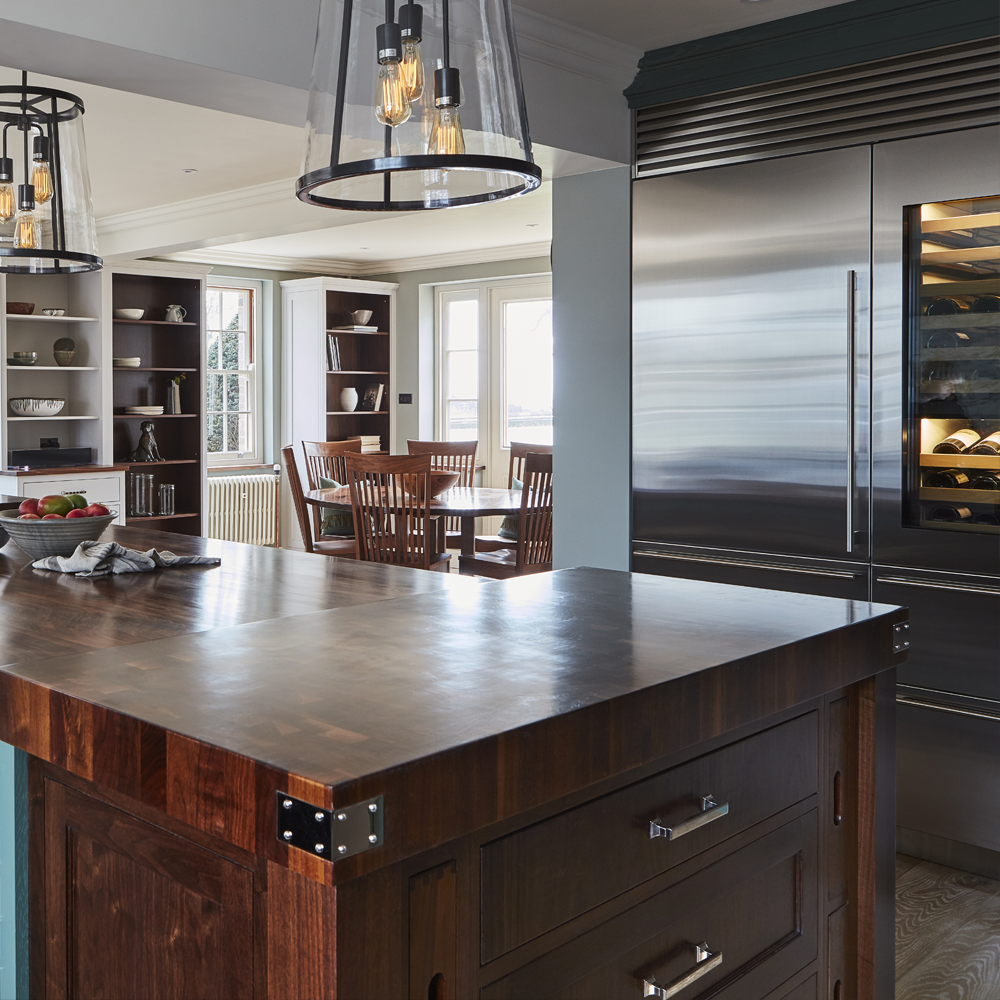 Modern kitchen with a dark wooden island, hanging glass pendant lights, stainless steel refrigerator, built-in wine cooler, and a dining area with wooden chairs in the background by large windows.