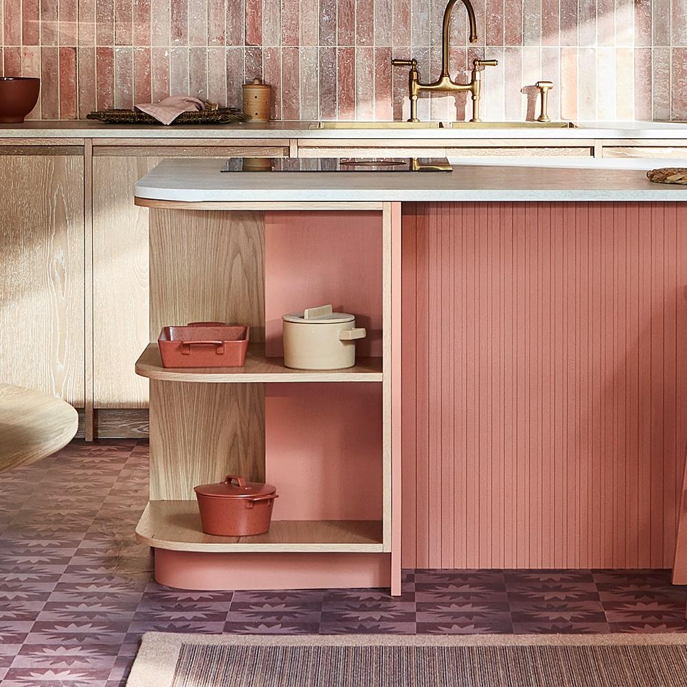 A modern kitchen features pink and beige tones, with a pink-tiled backsplash, light wood cabinets, and an island with open shelves holding ceramic cookware in pink and cream colors.