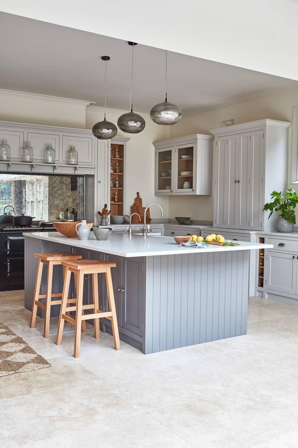 A bright kitchen with a large gray island, topped with a white countertop, lemons, and decorative items. Wooden stools are tucked under the island. Cabinets and a window are in the background. Three pendant lights hang above.