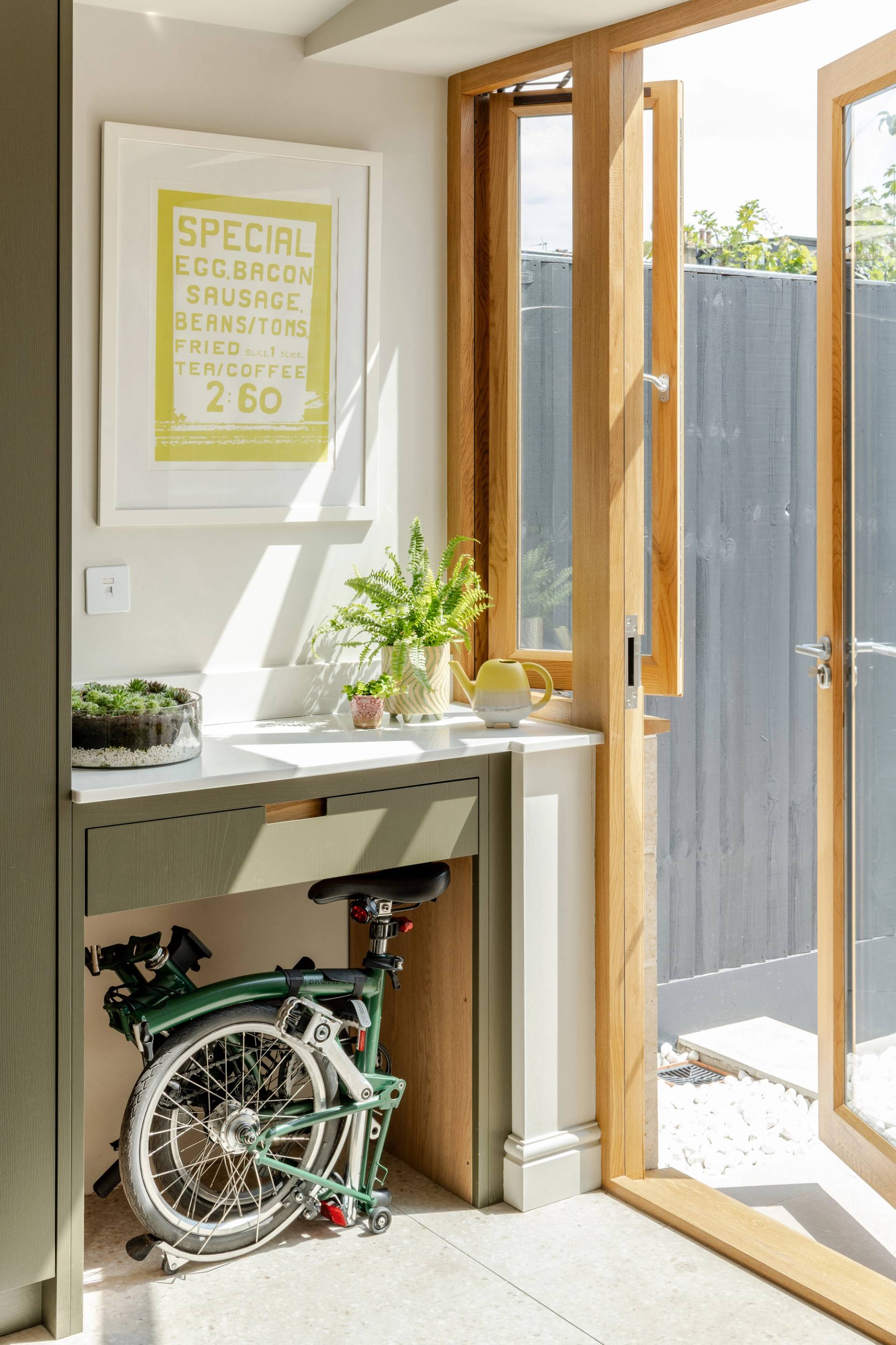 A modern drink station with olive-green cabinets, a sink, a mini fridge stocked with drinks, a coffee machine, shelves with liquor bottles, and a sign reading “CIN CIN!” above. A mirror reflects part of the kitchen.