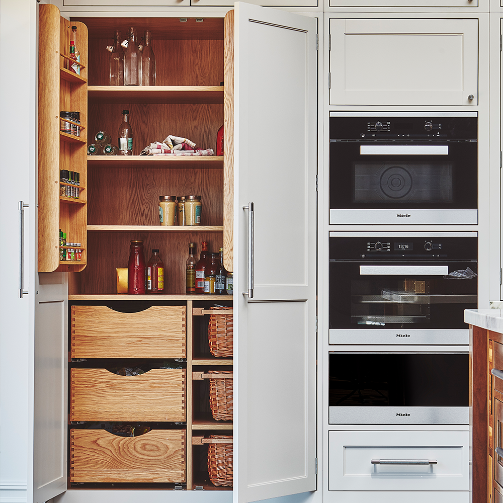 A neatly organized kitchen pantry with open doors, wooden shelves holding bottles, jars, and baskets, and built-in drawers below. Adjacent are modern built-in ovens and a microwave in white cabinetry.
