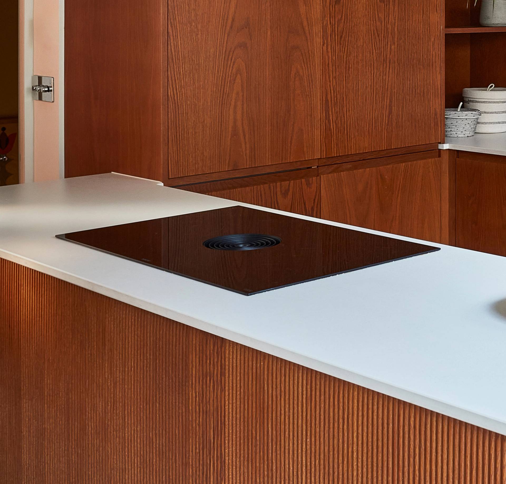 A modern kitchen featuring a sleek, black cooktop built into a white countertop. The cabinetry is made of wood with a vertical grain pattern, and there is a stainless steel door handle visible in the background.