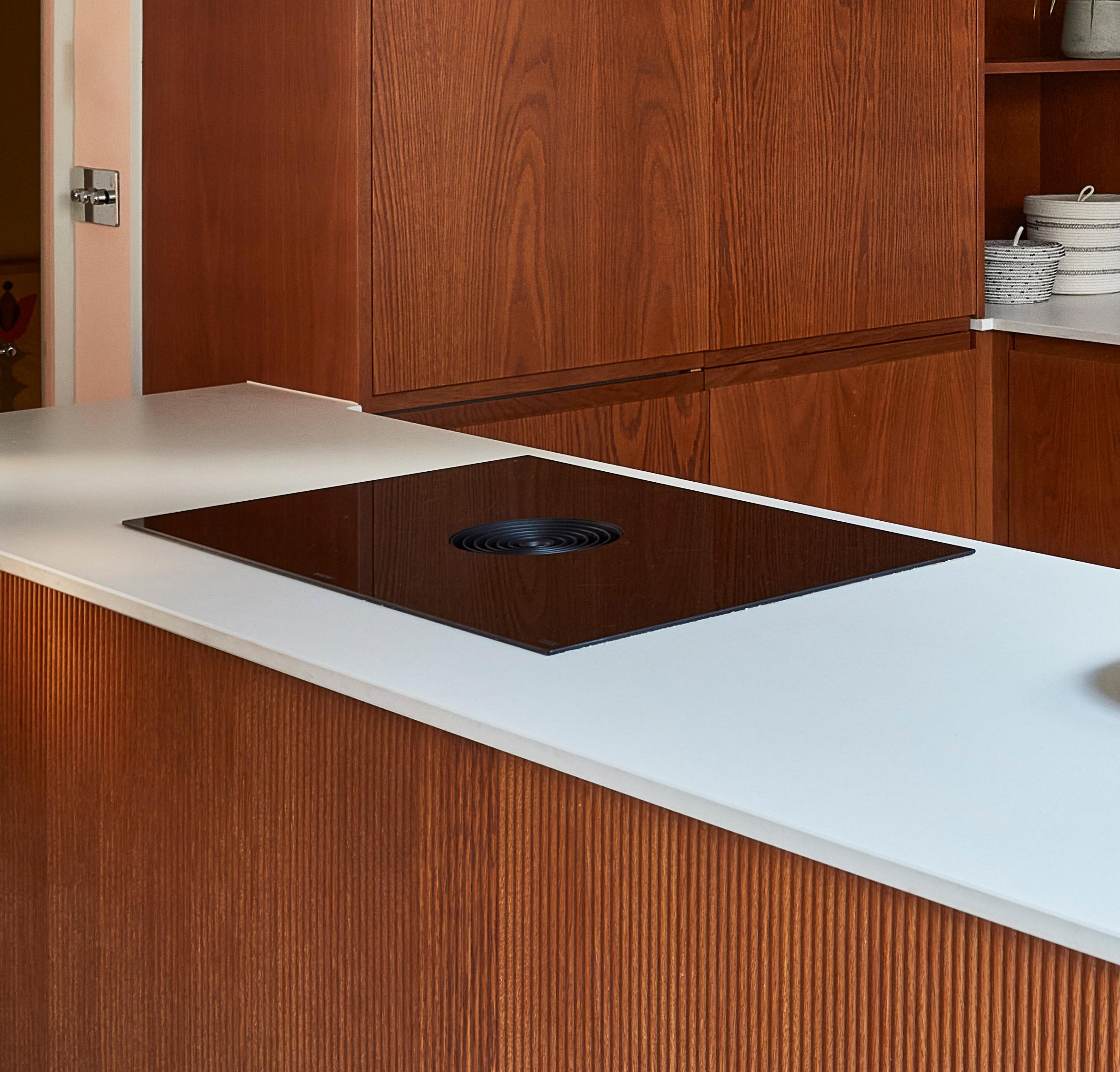 A modern kitchen featuring a sleek, black cooktop built into a white countertop. The cabinetry is made of wood with a vertical grain pattern, and there is a stainless steel door handle visible in the background.