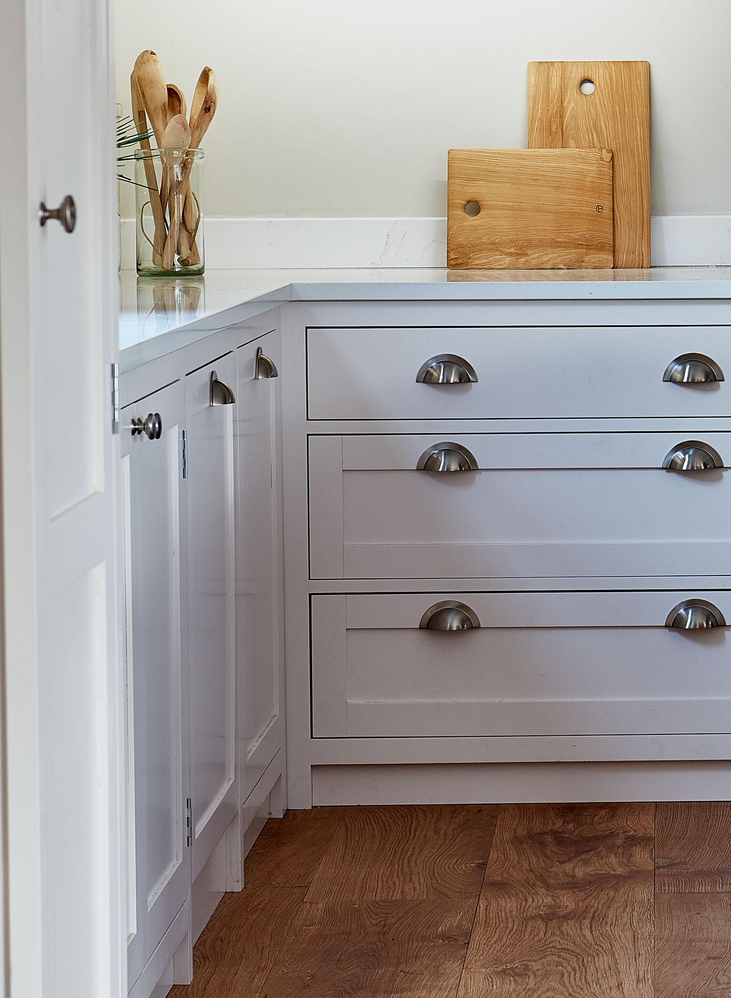 A corner of a modern kitchen with white cabinets and drawers, silver cup handles, a wood floor, two wooden cutting boards, and a glass jar holding wooden utensils on the countertop.