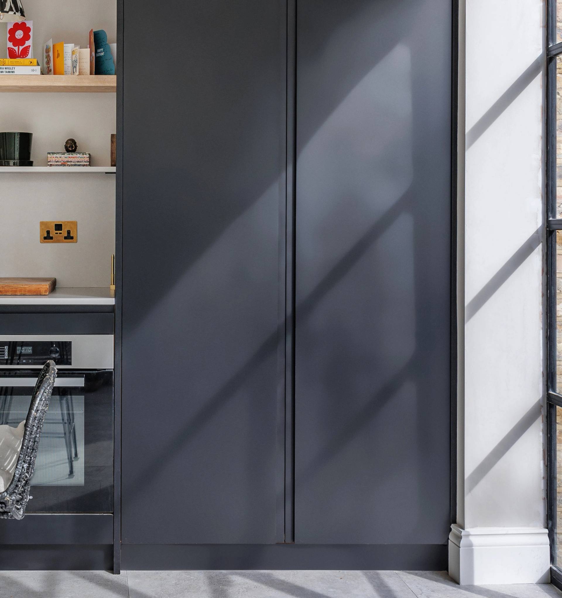 A modern interior with a dark gray built-in cabinet next to shelves holding books and decor. Sunlight streams through a large window, casting shadows on the wall and cabinet.