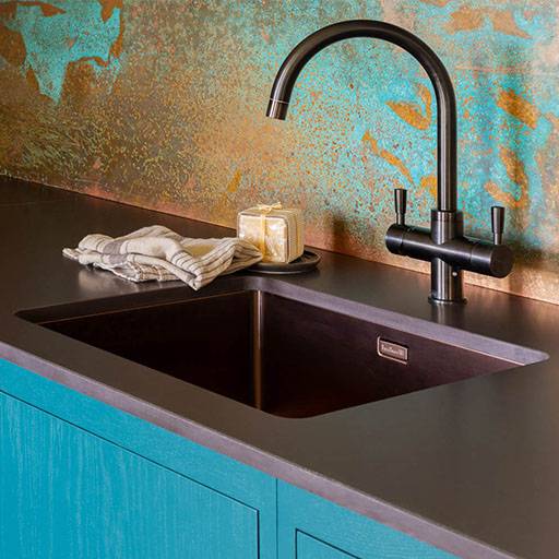 A modern kitchen sink with a matte black faucet, dark countertop, turquoise cabinets, a striped dish towel, and a bar of soap in a holder. The backsplash features a textured blue and copper pattern.