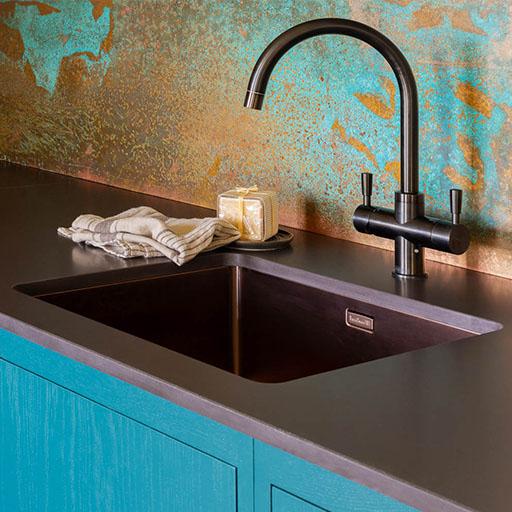 A modern kitchen sink with a matte black faucet, dark countertop, turquoise cabinets, a striped dish towel, and a bar of soap in a holder. The backsplash features a textured blue and copper pattern.
