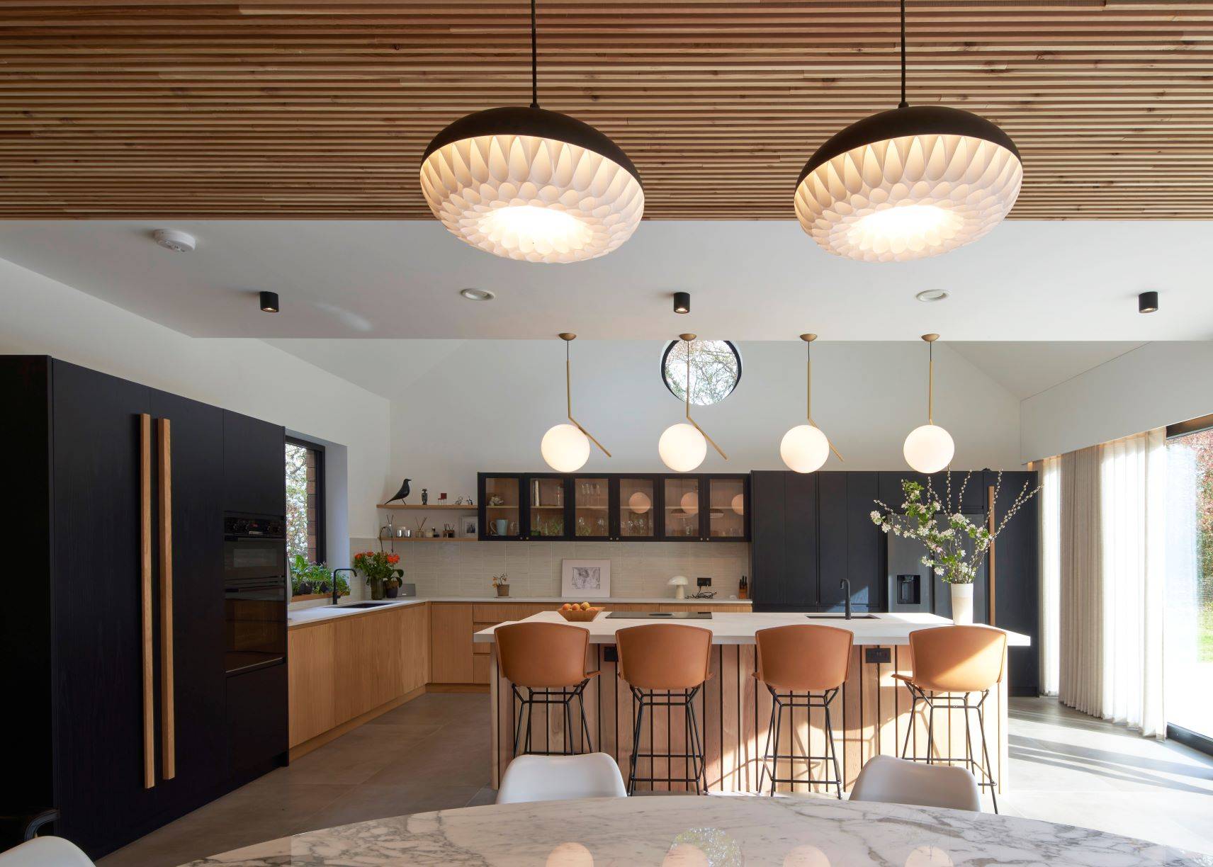 Modern kitchen with a large marble island, surrounded by tan upholstered bar stools. Black cabinets contrast with natural wood finishes. Unique hanging lights illuminate the space, and a row of windows allows natural daylight to enter.