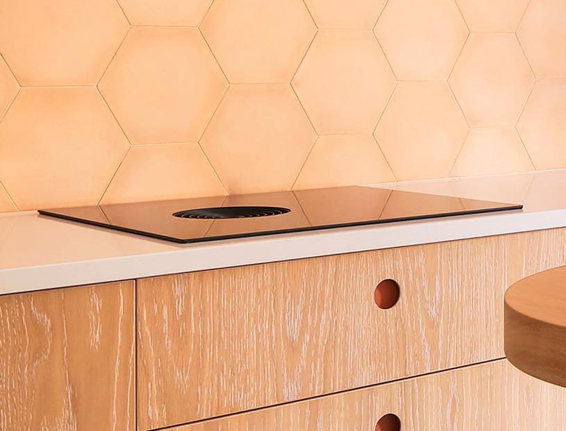 A modern kitchen with light wood cabinets, a white countertop, and a built-in black induction cooktop with an integrated vent, set against a peach hexagon tile backsplash.
