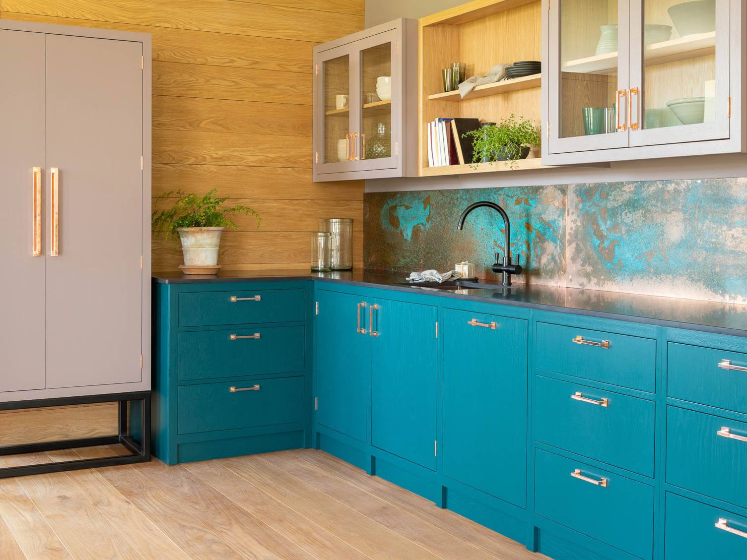 A modern kitchen with teal lower cabinets, a black countertop, and a copper backsplash. Upper cabinets are beige with glass doors. A large fridge stands to the left. There's a potted plant and various dishware on the shelves. Floors are light wood.