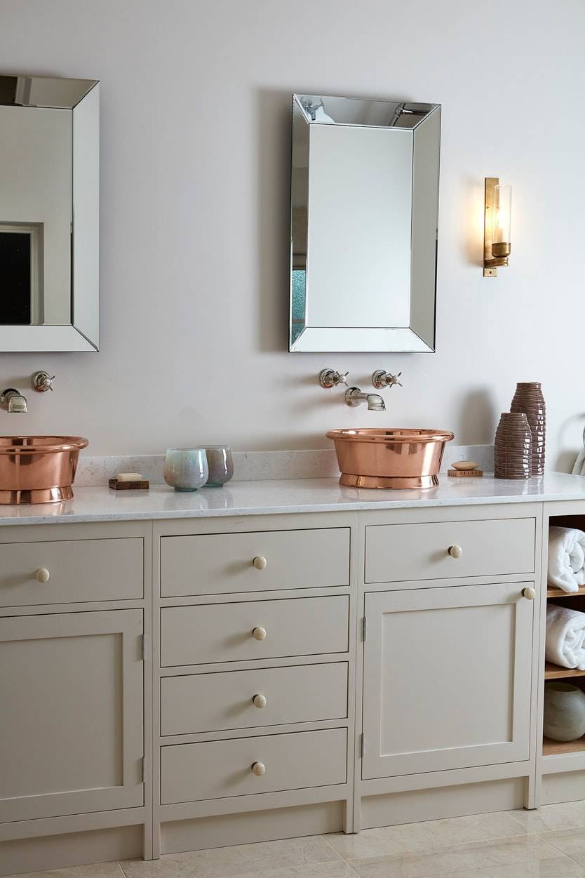 A modern bathroom with cream cabinets, a countertop holding a shiny copper basin sink, a wall-mounted faucet, a square mirror, a wall sconce, and shelves with rolled white towels and decorative items.