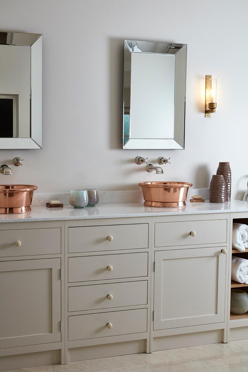 A modern bathroom with cream cabinets, a countertop holding a shiny copper basin sink, a wall-mounted faucet, a square mirror, a wall sconce, and shelves with rolled white towels and decorative items.