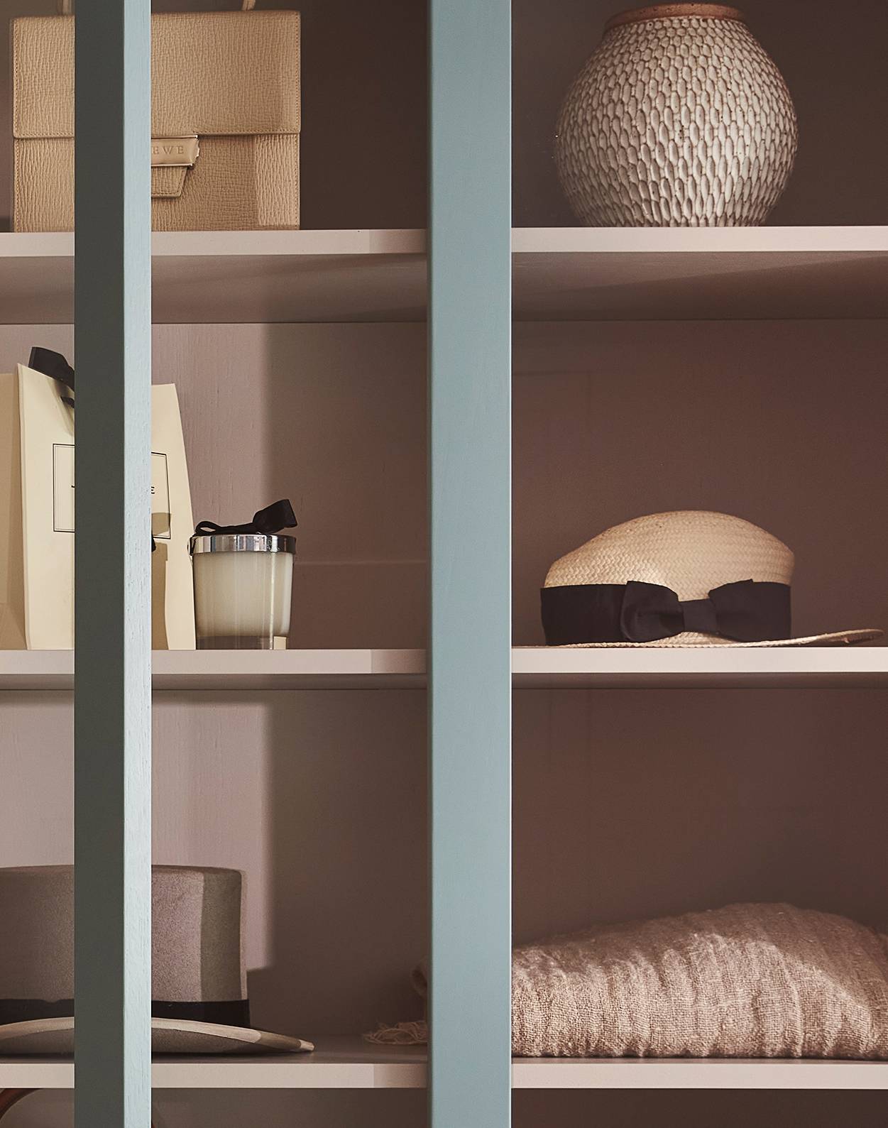 Shelves with decorative items including stacked boxes, a textured vase, a candle, a folded sweater, and hats, viewed partially through vertical blue slats