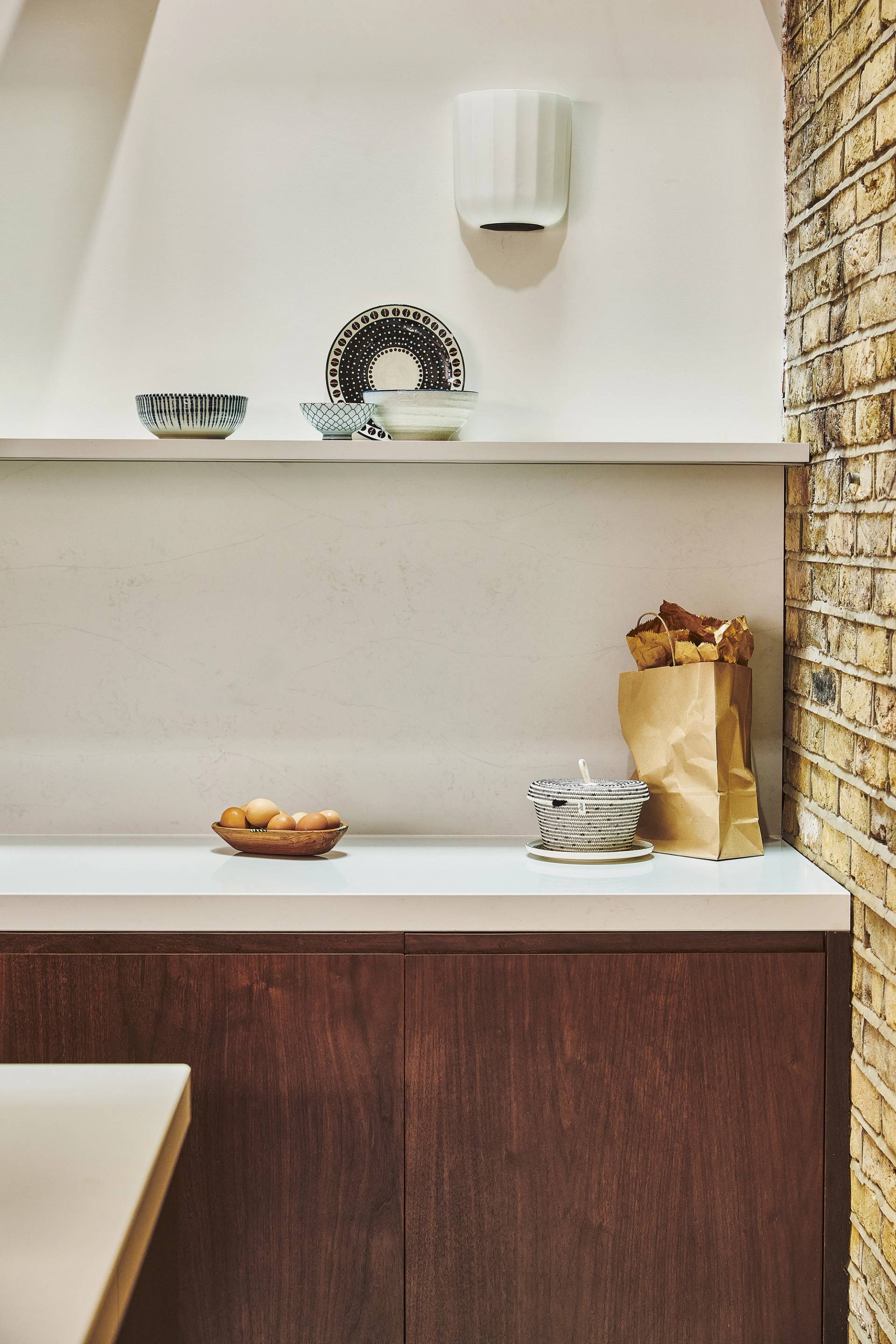 A modern kitchen with wooden cabinets, a brick wall, a white shelf holding patterned bowls and plates, a brown paper bag with baguettes, a bowl of eggs, and a gray container on the countertop.