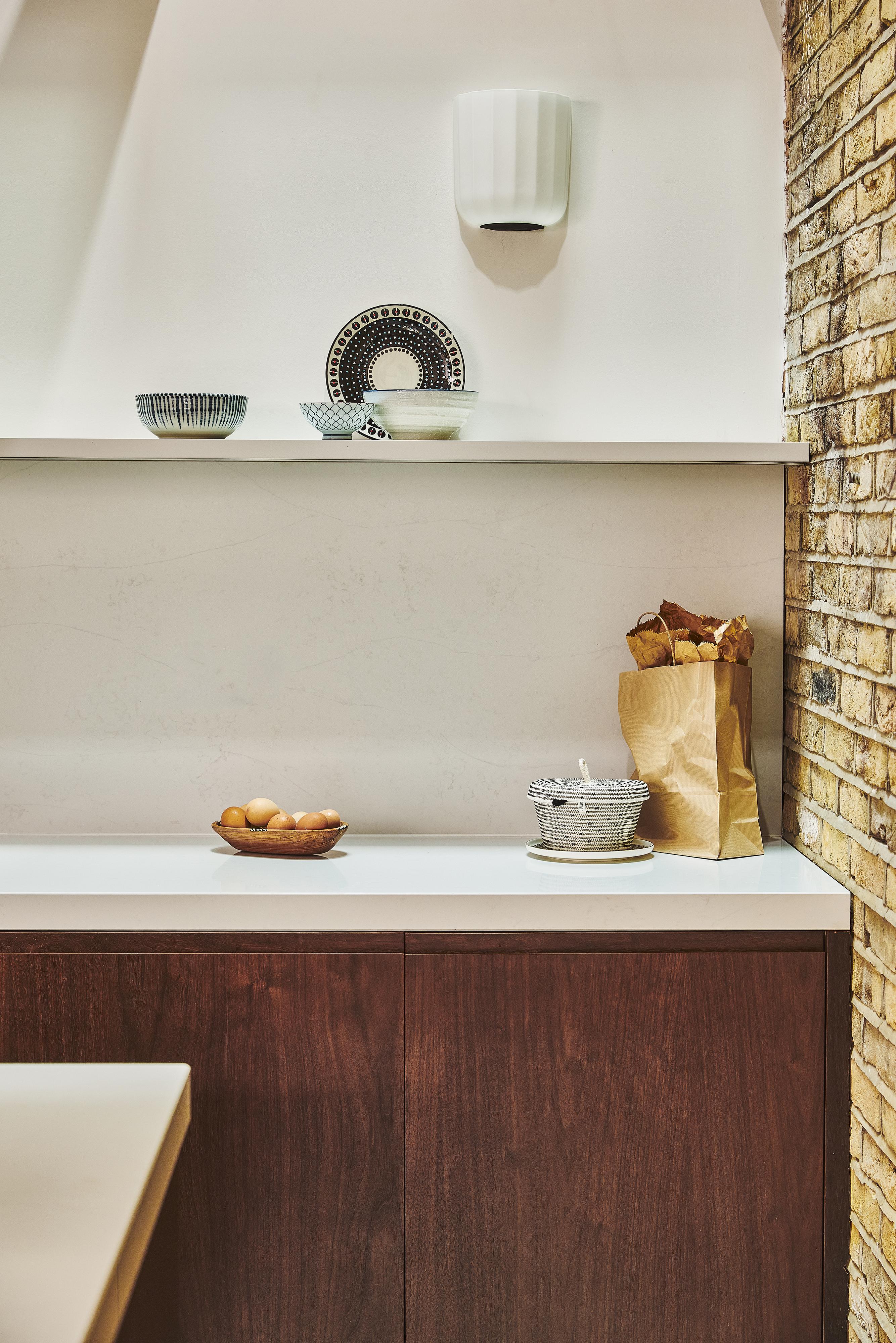 A modern kitchen with wooden cabinets, a brick wall, a white shelf holding patterned bowls and plates, a brown paper bag with baguettes, a bowl of eggs, and a gray container on the countertop.